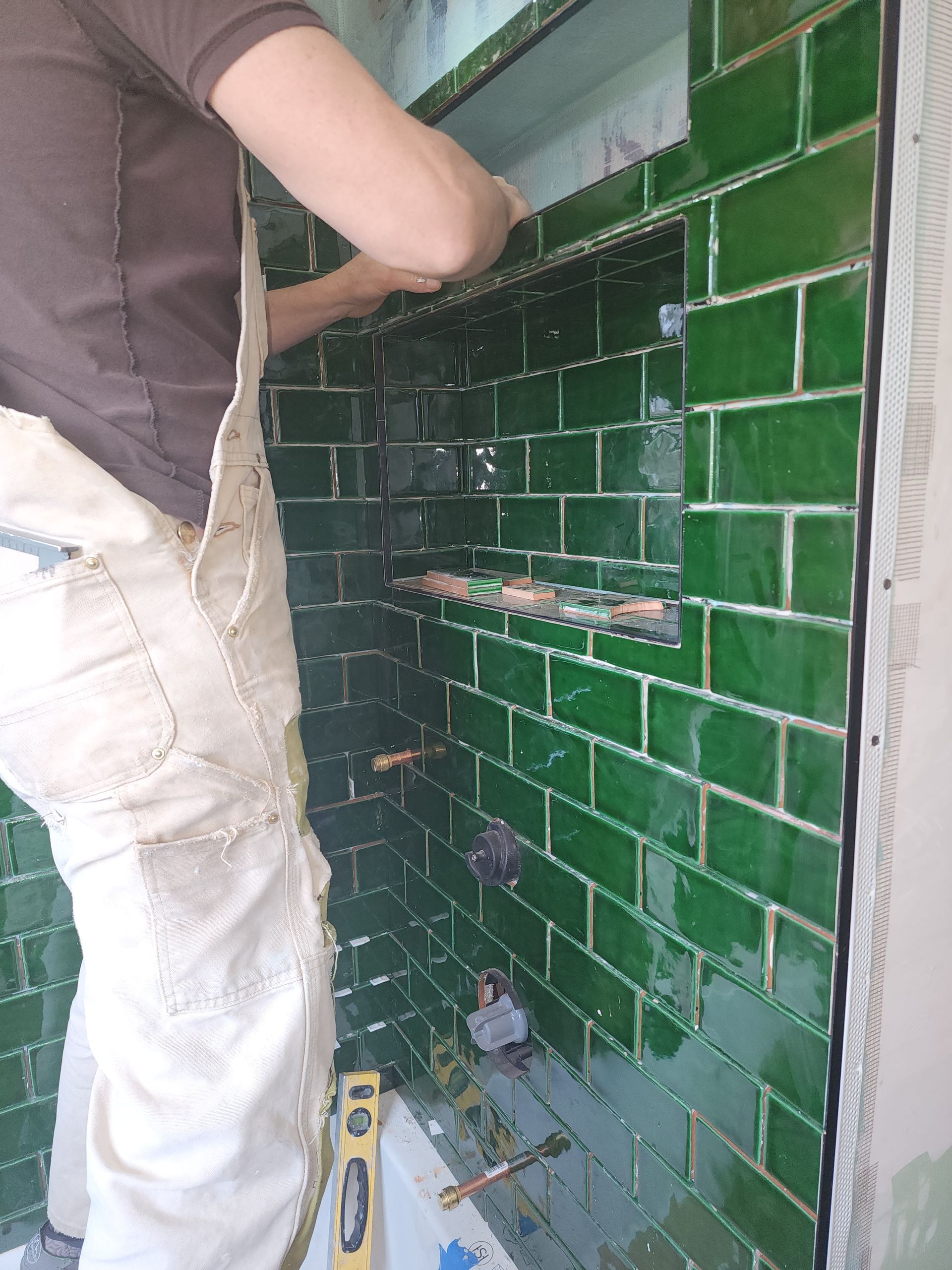 A person wearing white overalls installs glossy green subway tiles on a shower wall with a built-in recessed shelf.