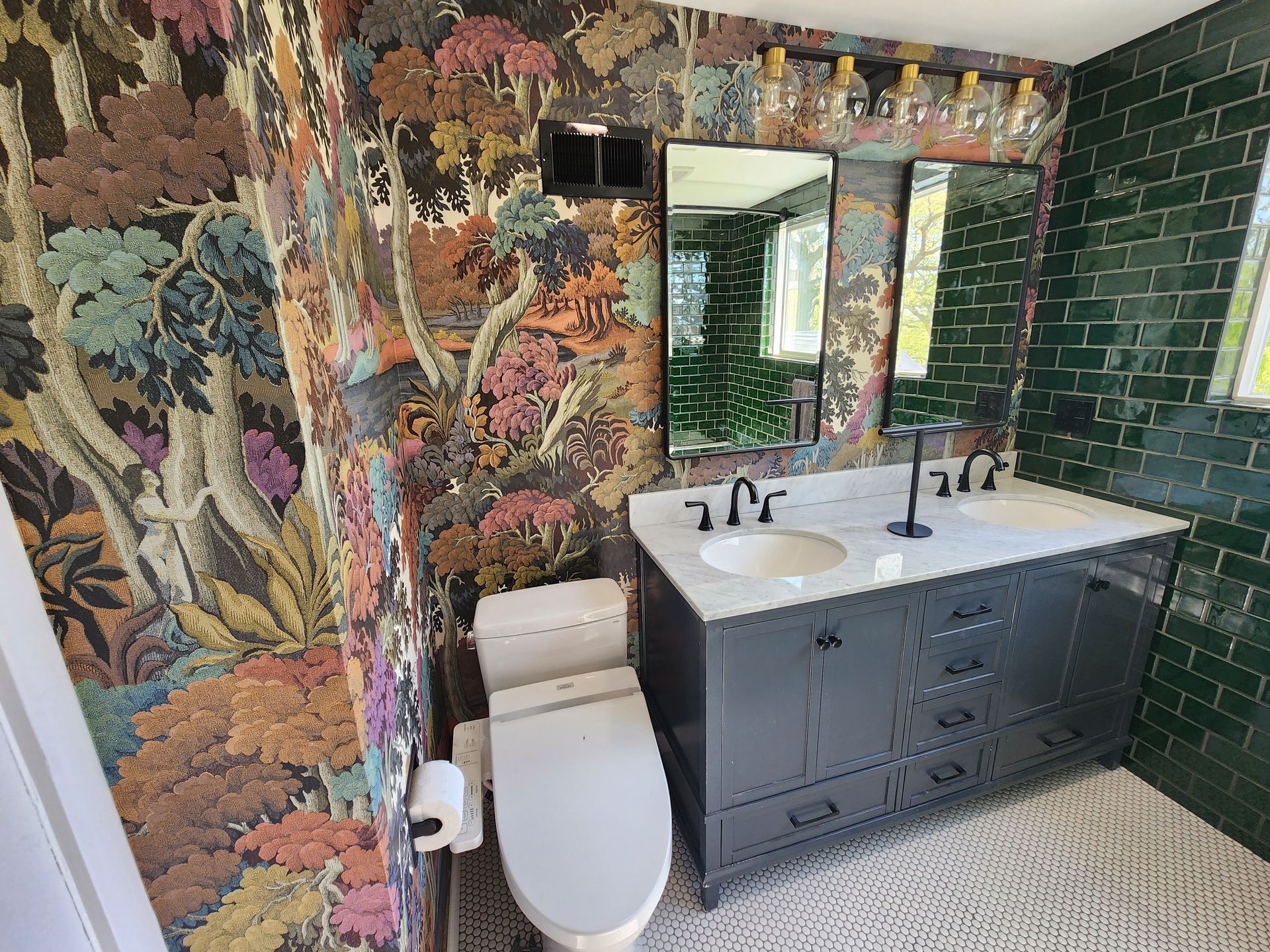 A bathroom with floral wallpaper, a dark grey vanity with two sinks, a white toilet, and dark green subway tile walls.