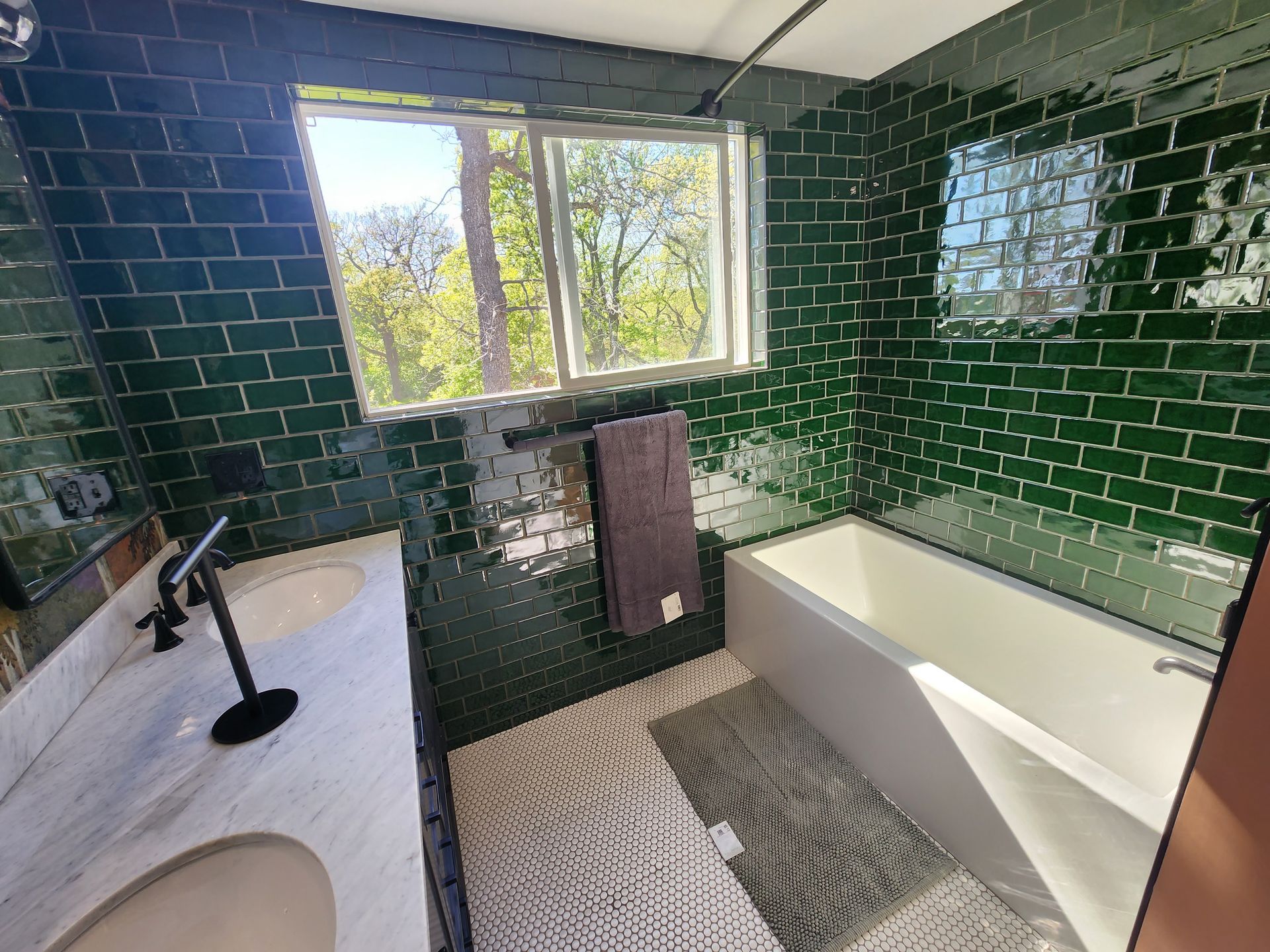 A bathroom with dark green subway tile walls, a white double-sink marble vanity, and a white bathtub under a window.