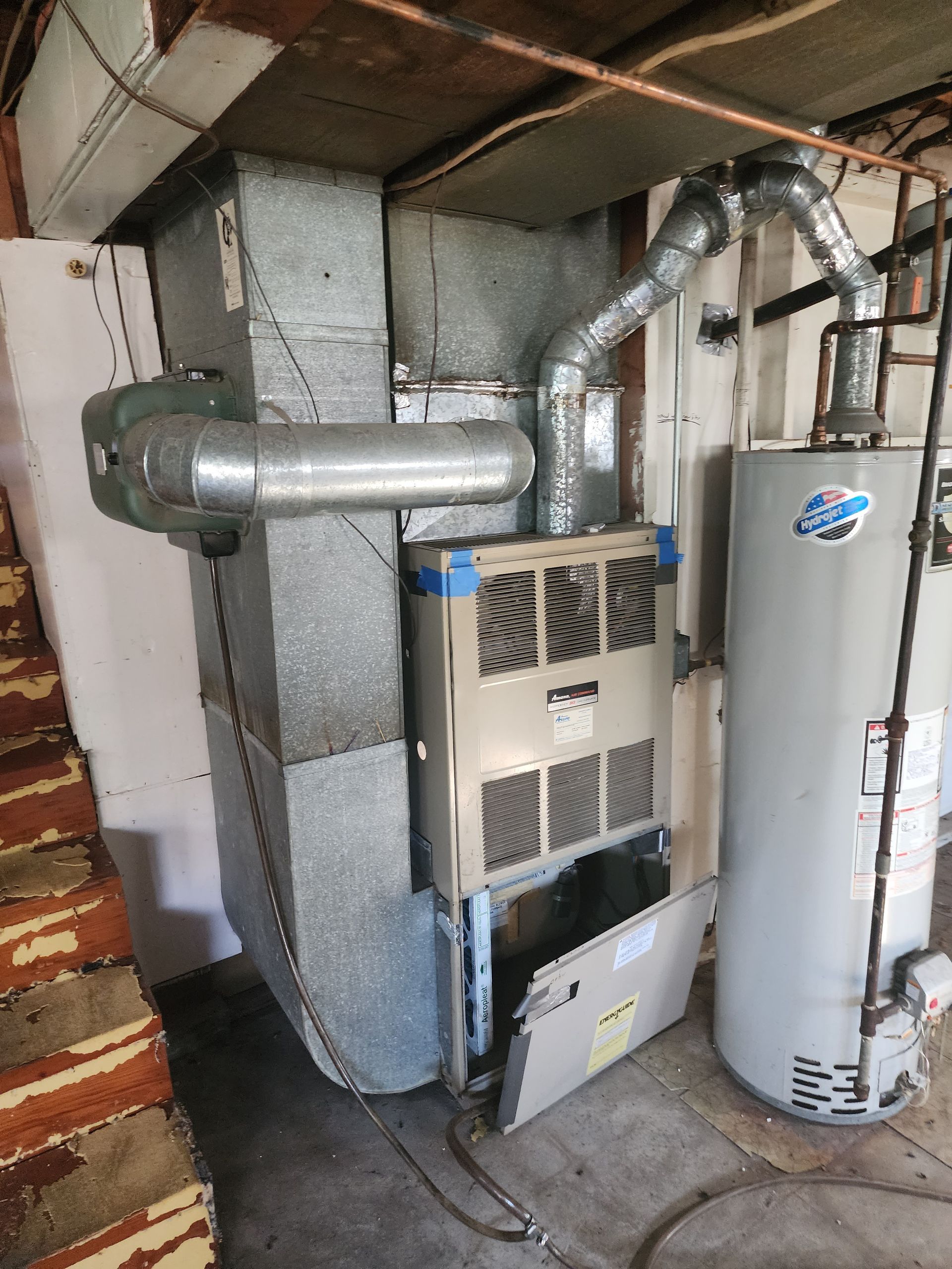 An older gas furnace and a water heater stand in a basement with an open metal access panel on the furnace.