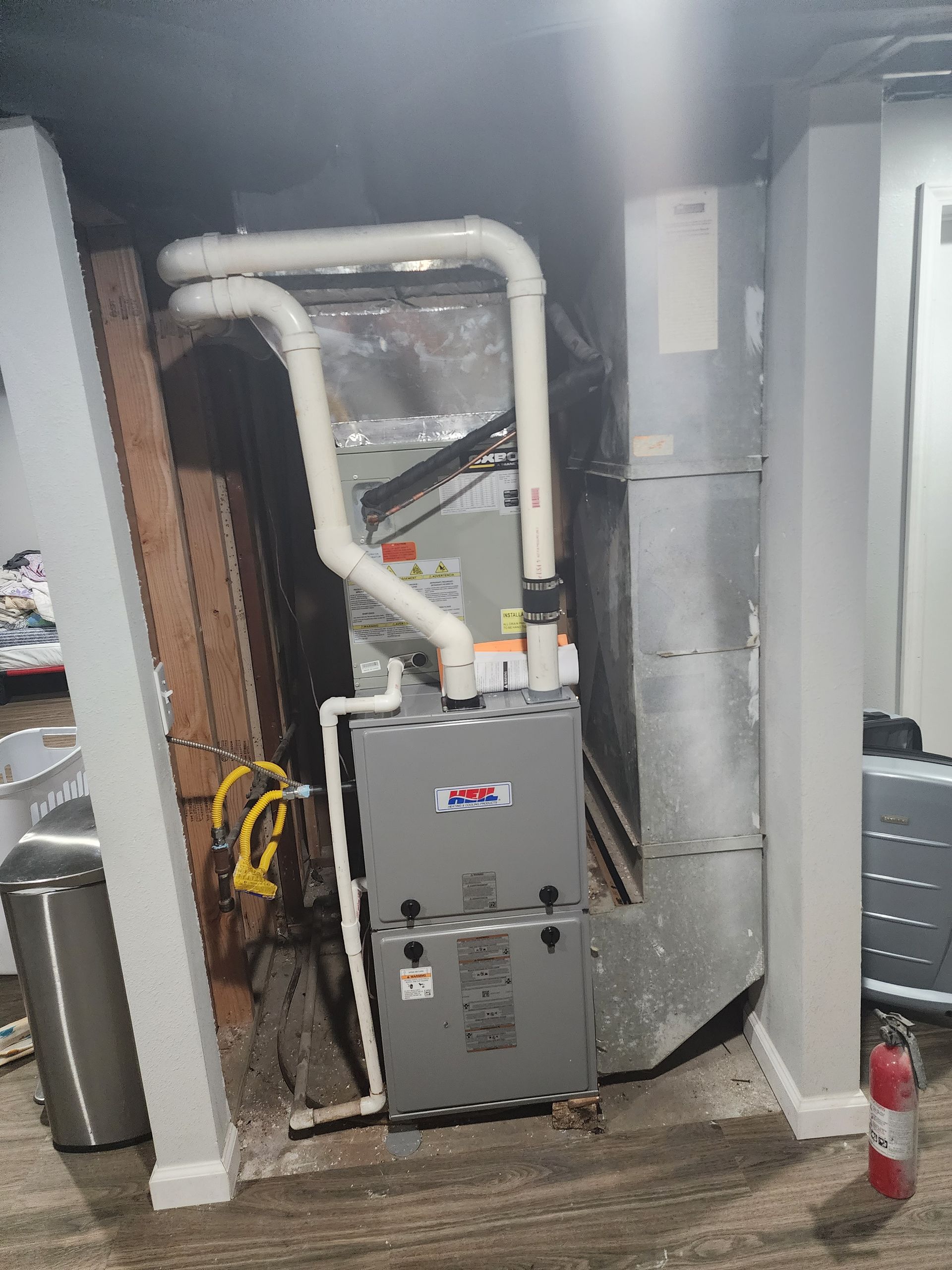 A grey gas furnace with white PVC venting pipes, standing in a basement corner next to a silver metal air duct.