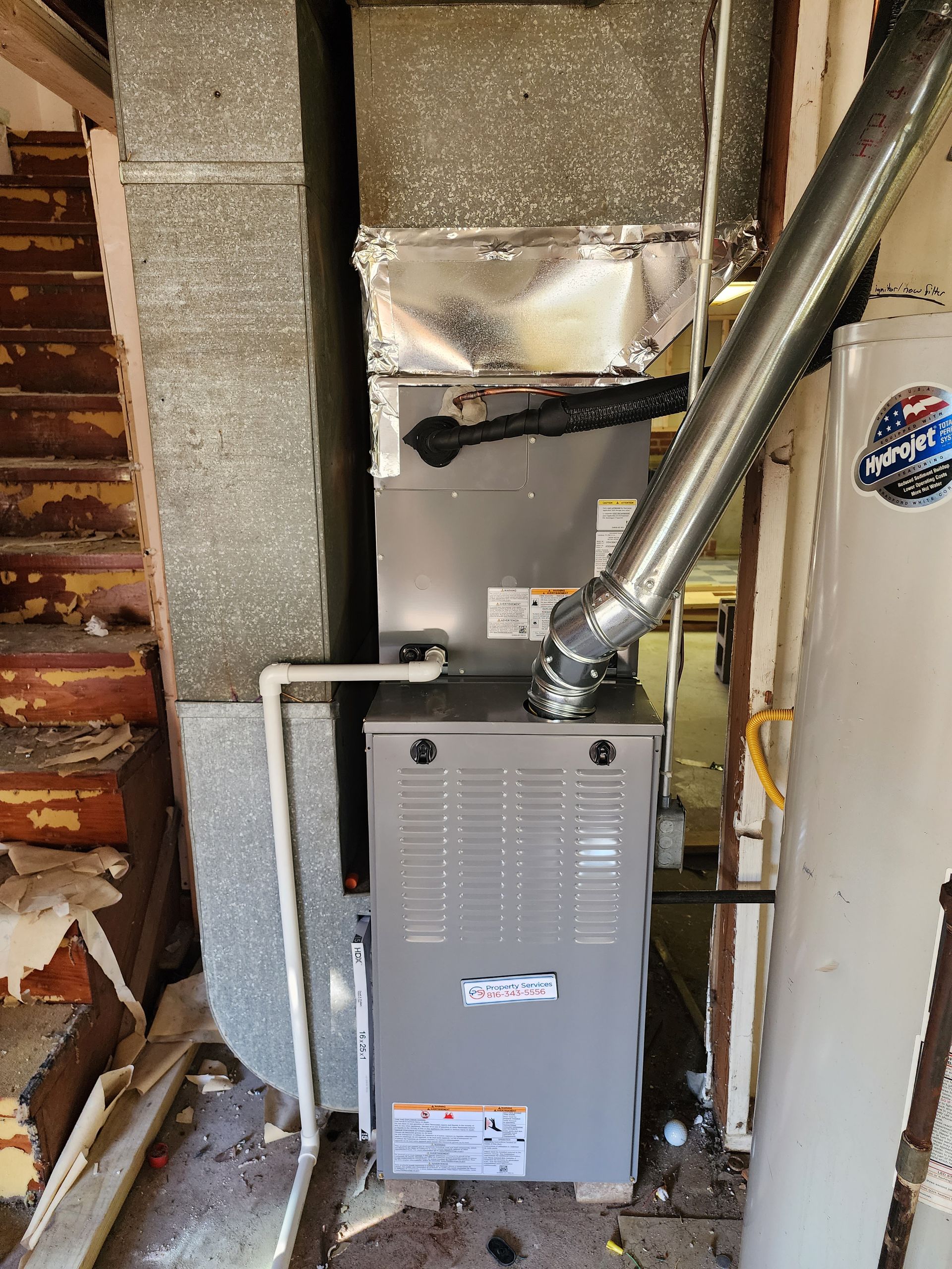 A gray furnace unit positioned in a basement next to a staircase and a water heater, with attached metal ductwork.