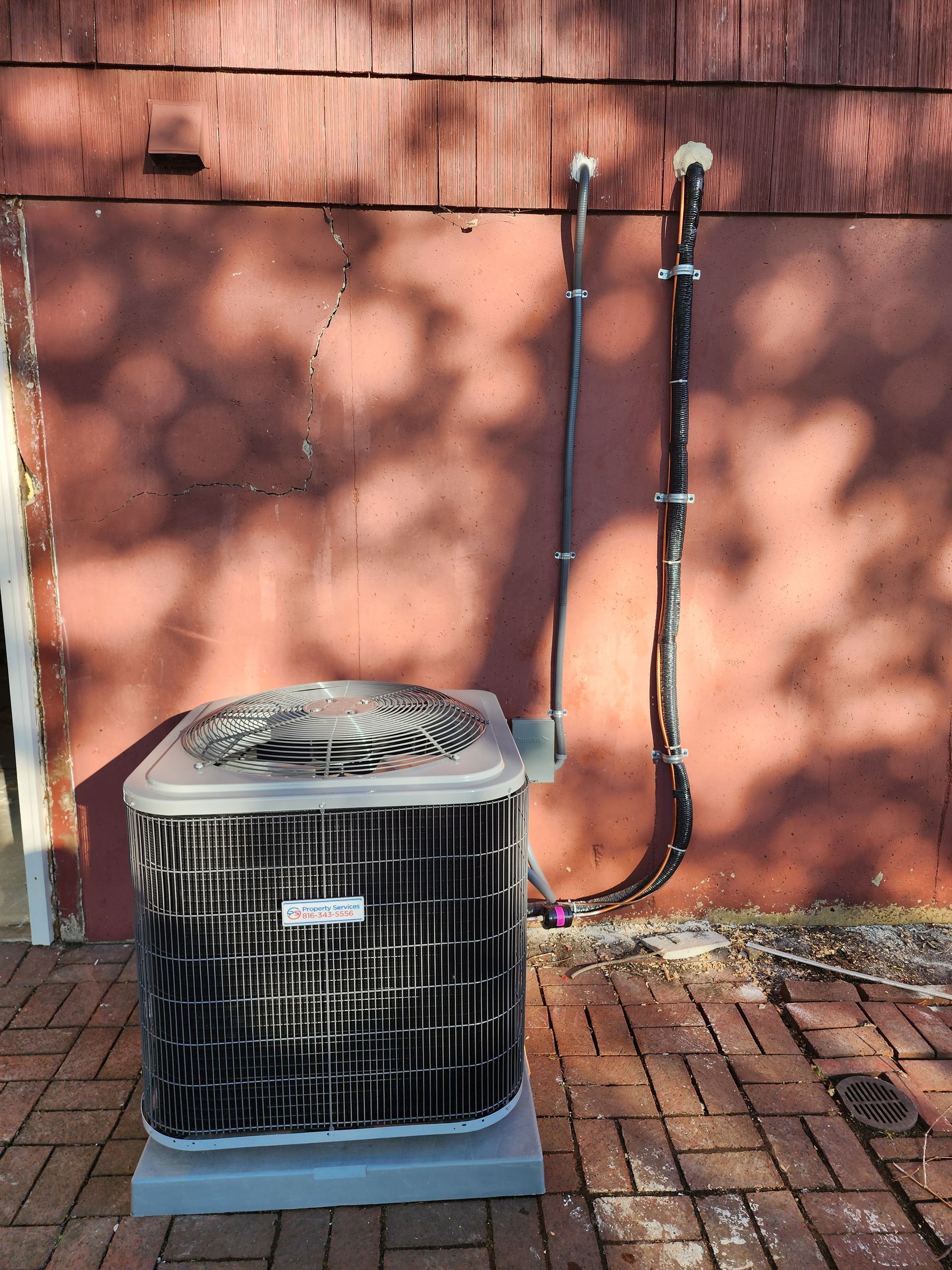 An outdoor air conditioning unit sits on a brick patio against a red wall, with electrical cables running up the side.
