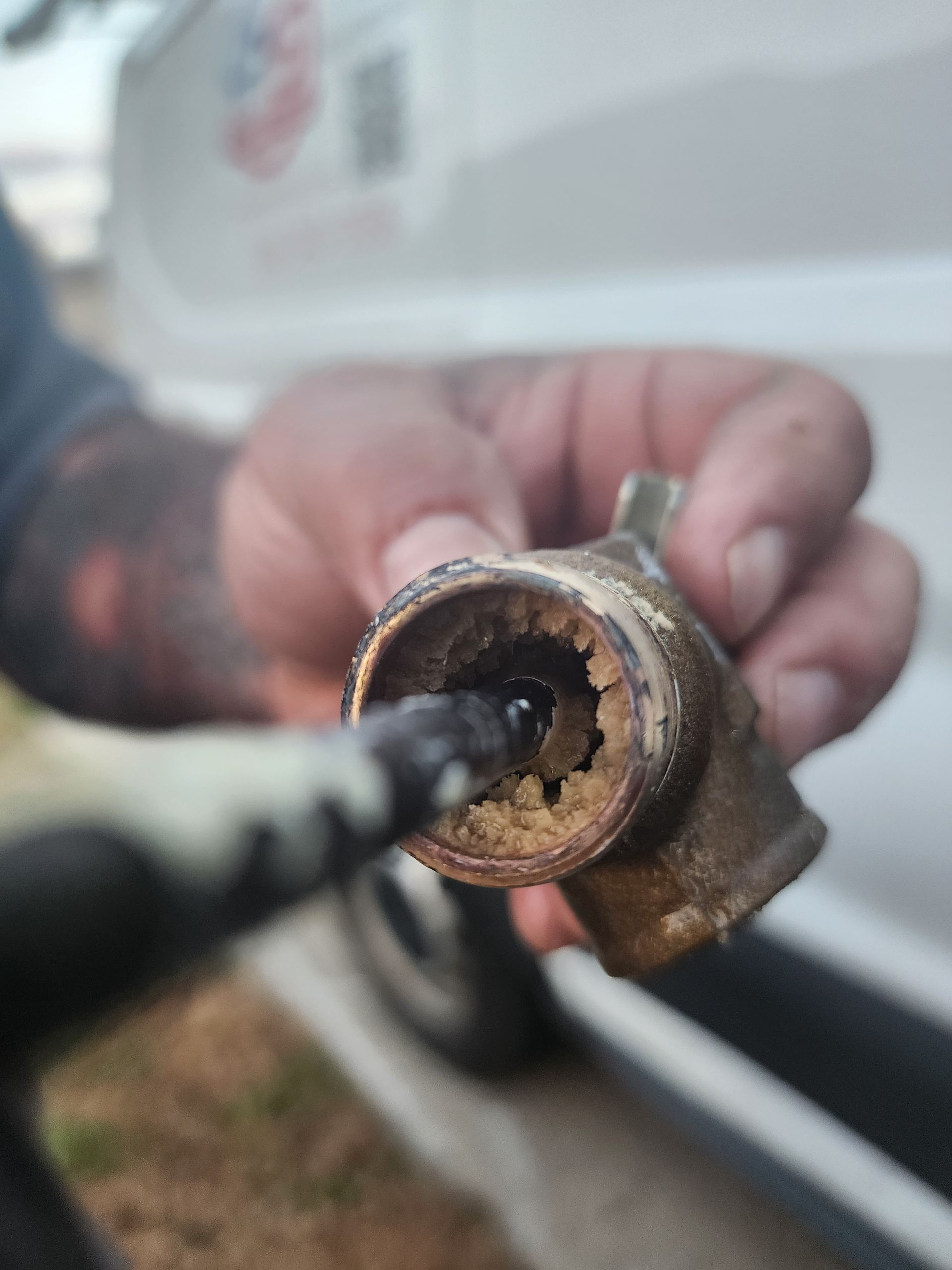 A hand holds a brass pipe fitting clogged with white mineral scale buildup, with a tool inserted into the opening.