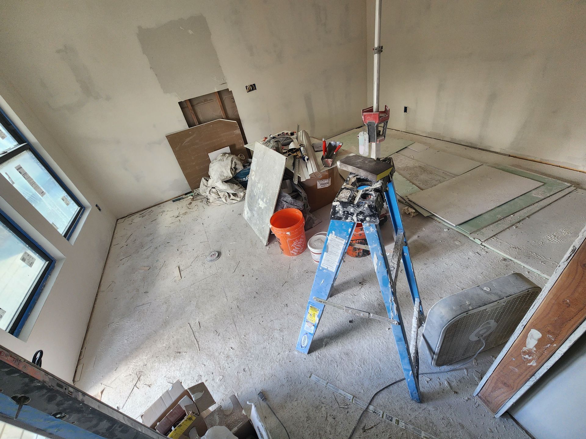A room under renovation with a blue stepladder, an orange bucket, and drywall materials on a dusty floor.