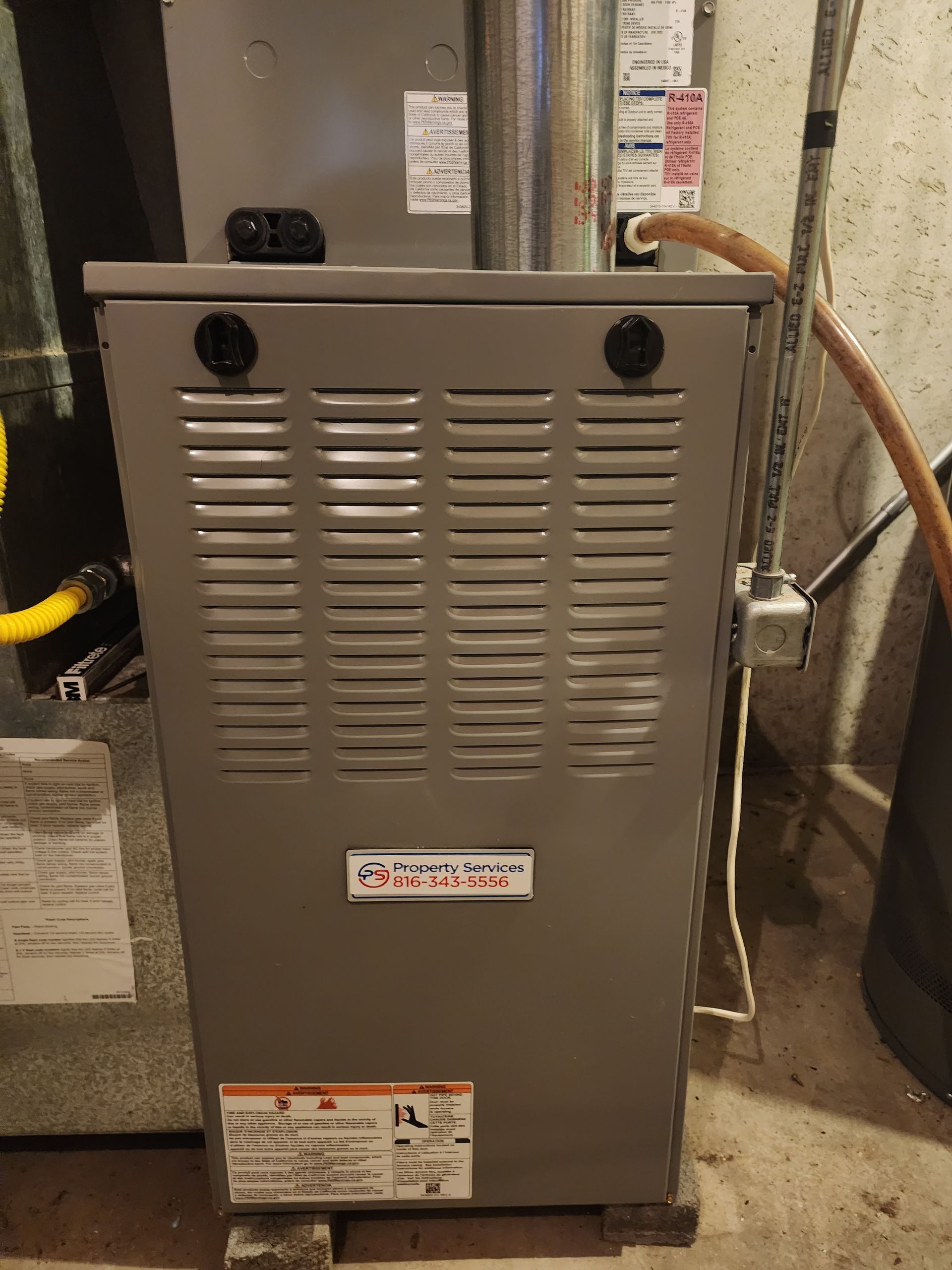 A grey vertical HVAC furnace unit positioned in a basement next to ductwork and utility connections.