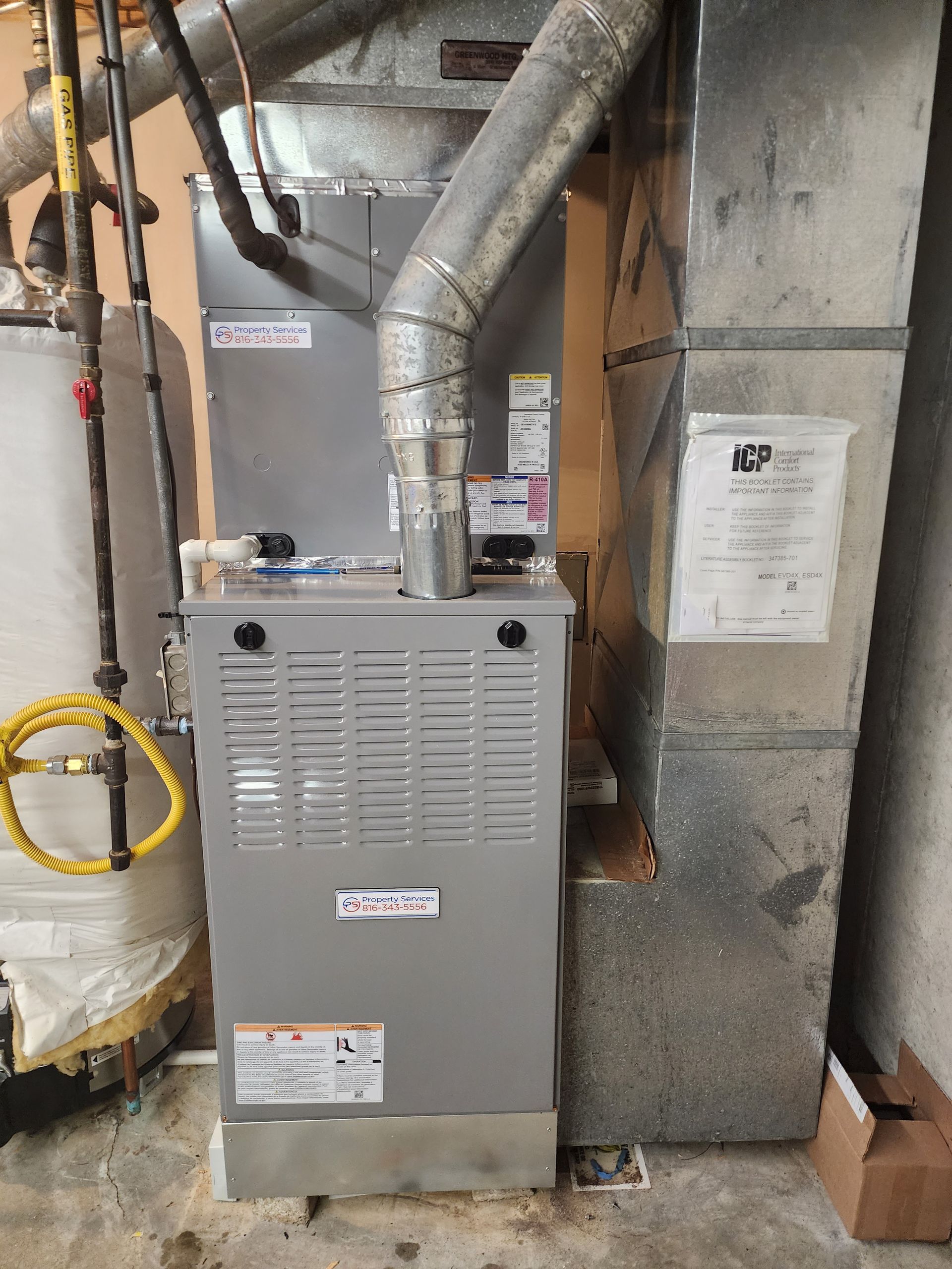 A grey HVAC furnace unit sits in a utility room next to a water heater and a metal ductwork stack.