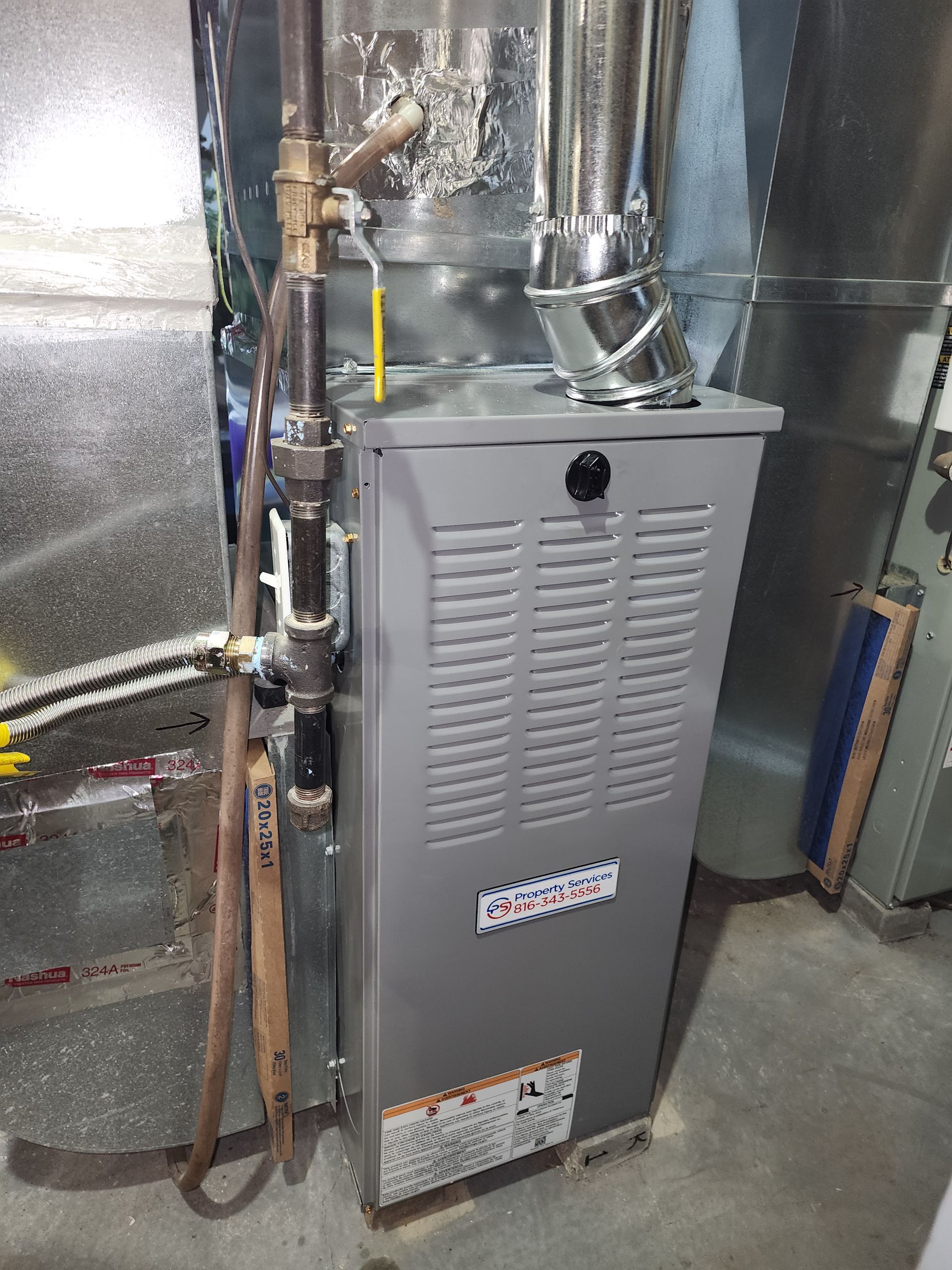 A vertical gray HVAC furnace unit with a metal exhaust pipe and gas line connections in a basement setting.