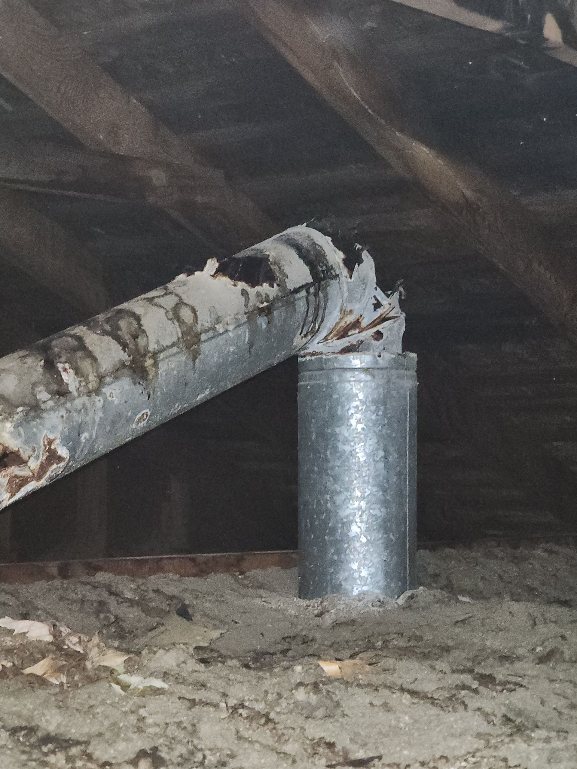 A detached metal pipe segment in a dusty attic, misaligned from the vertical vent stack below.