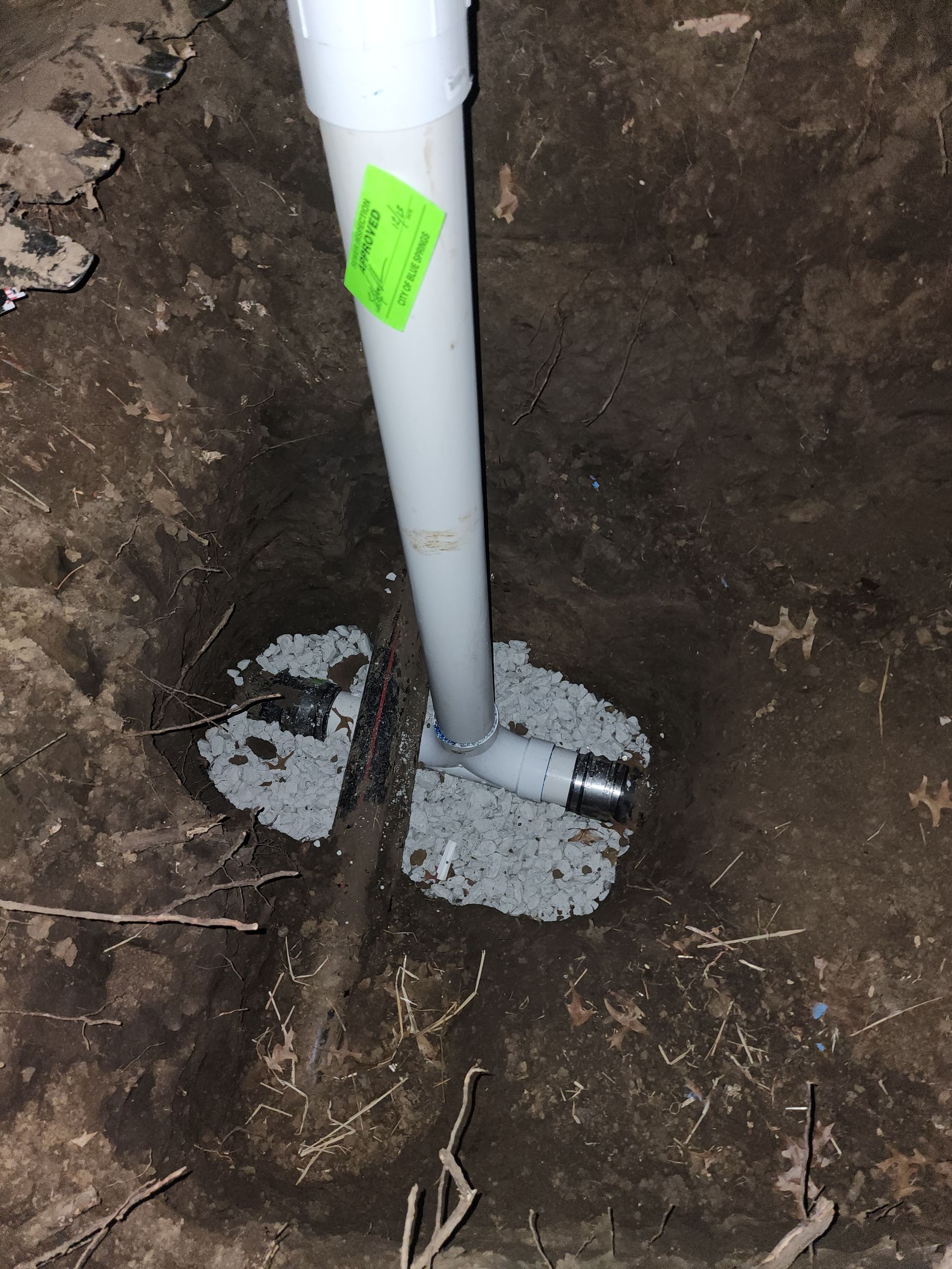 A vertical white PVC pipe stands in a hole in the ground, connected to a horizontal pipe set into concrete.