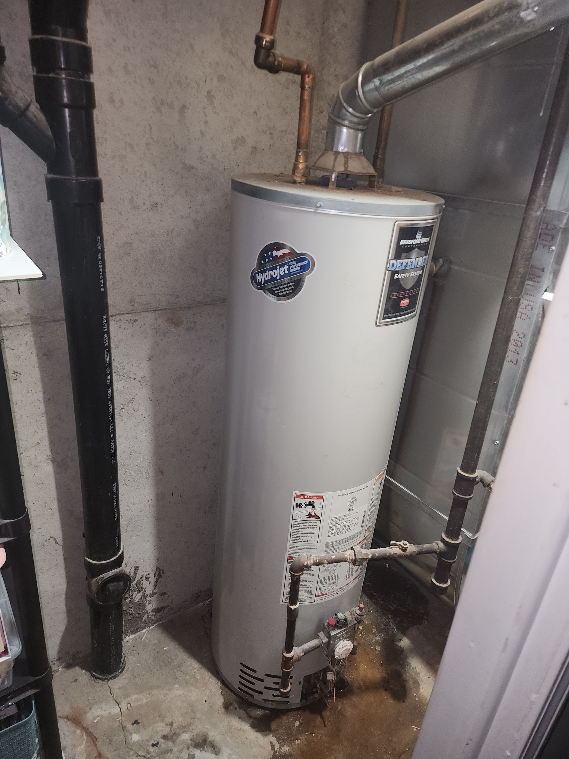 A tall, light-gray residential water heater standing in a utility room next to a black vertical drainage pipe.