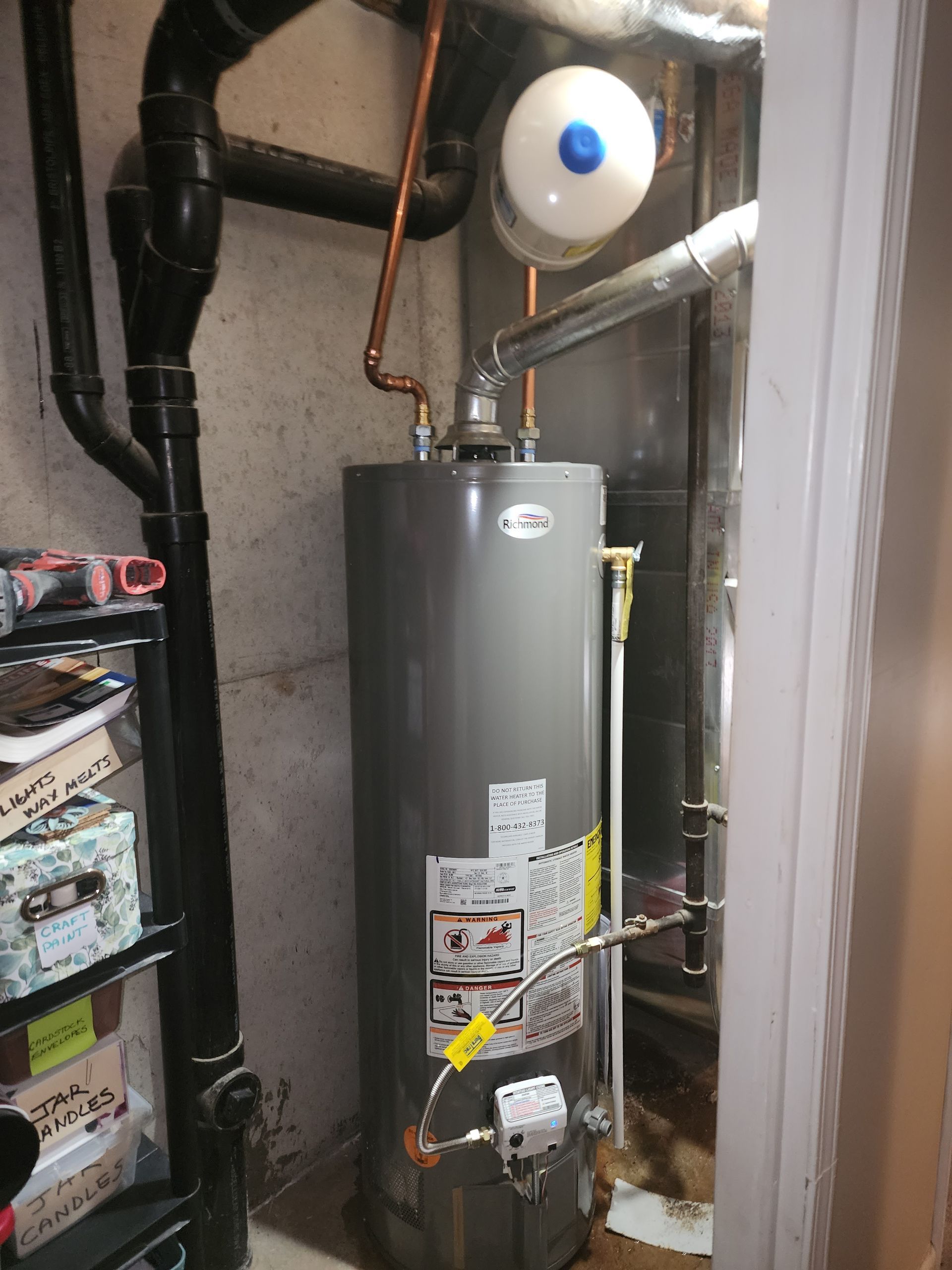 A gray residential water heater connected to pipes and a white expansion tank in a utility area.