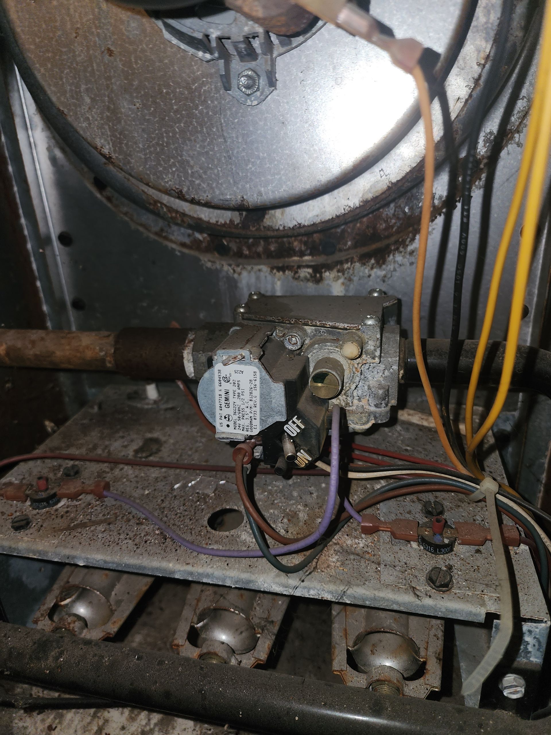A dusty, metallic gas valve and burner assembly inside an HVAC furnace unit, with visible wires and burner tubes.