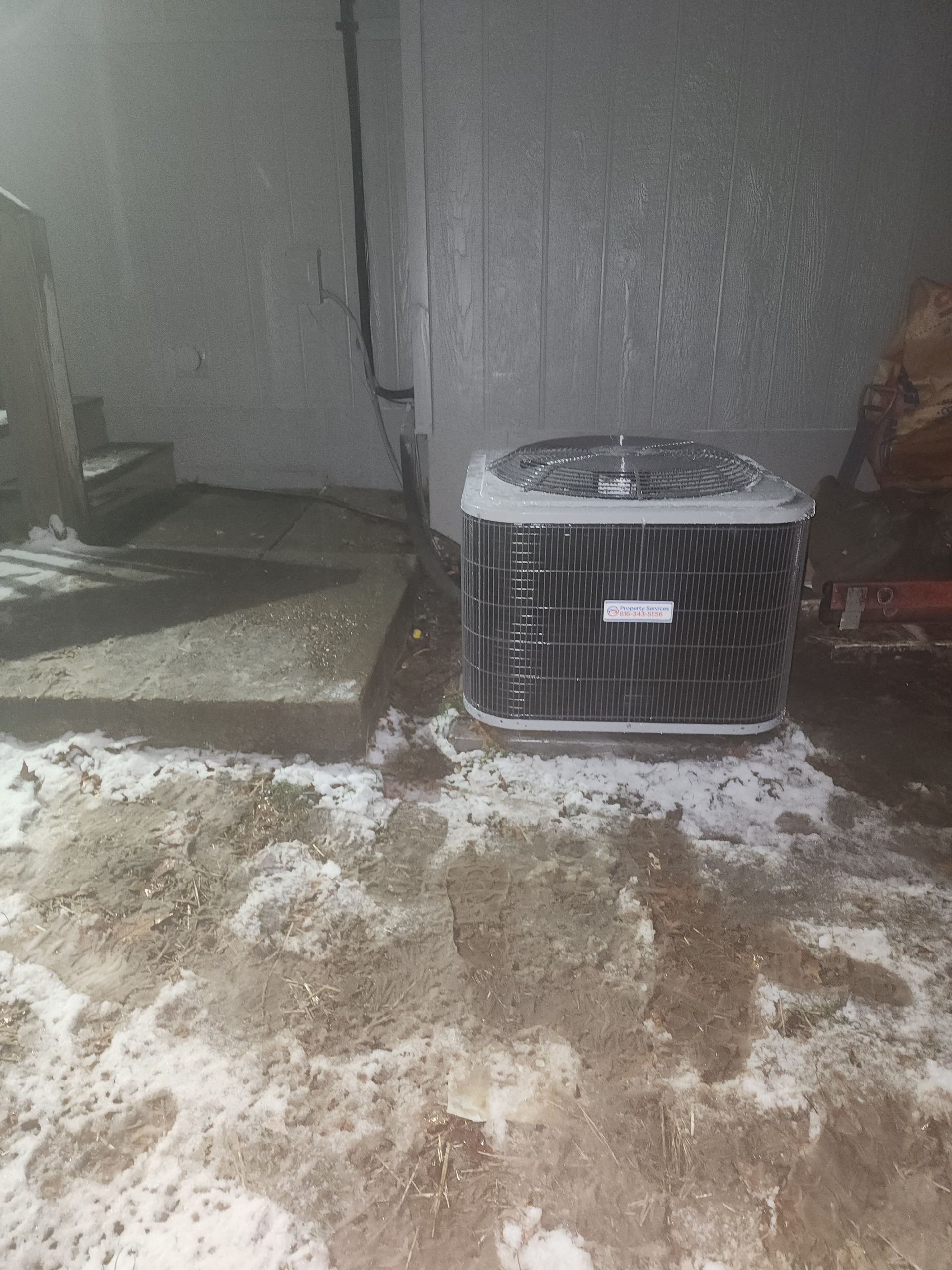 An outdoor air conditioning unit sits on the ground next to a concrete step, surrounded by patches of snow and dirt.
