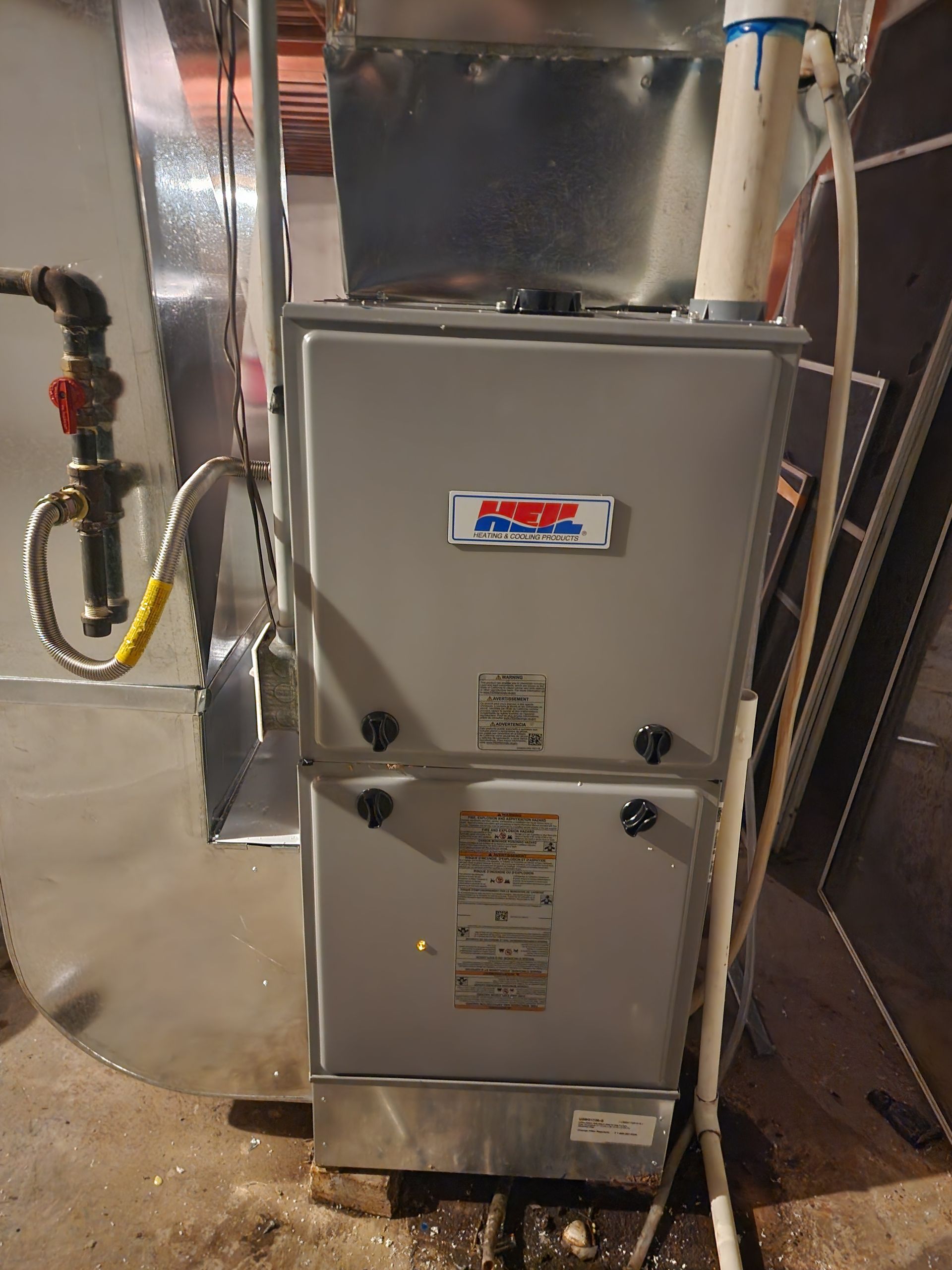 A gray, upright gas furnace installed in a utility space, connected to metal ductwork and white PVC exhaust piping.