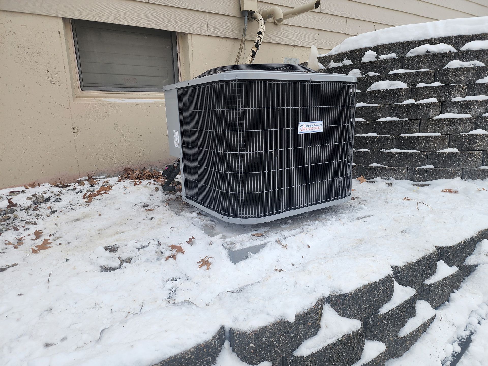 A residential HVAC outdoor unit sits on a concrete pad next to a stone retaining wall in a snowy yard.