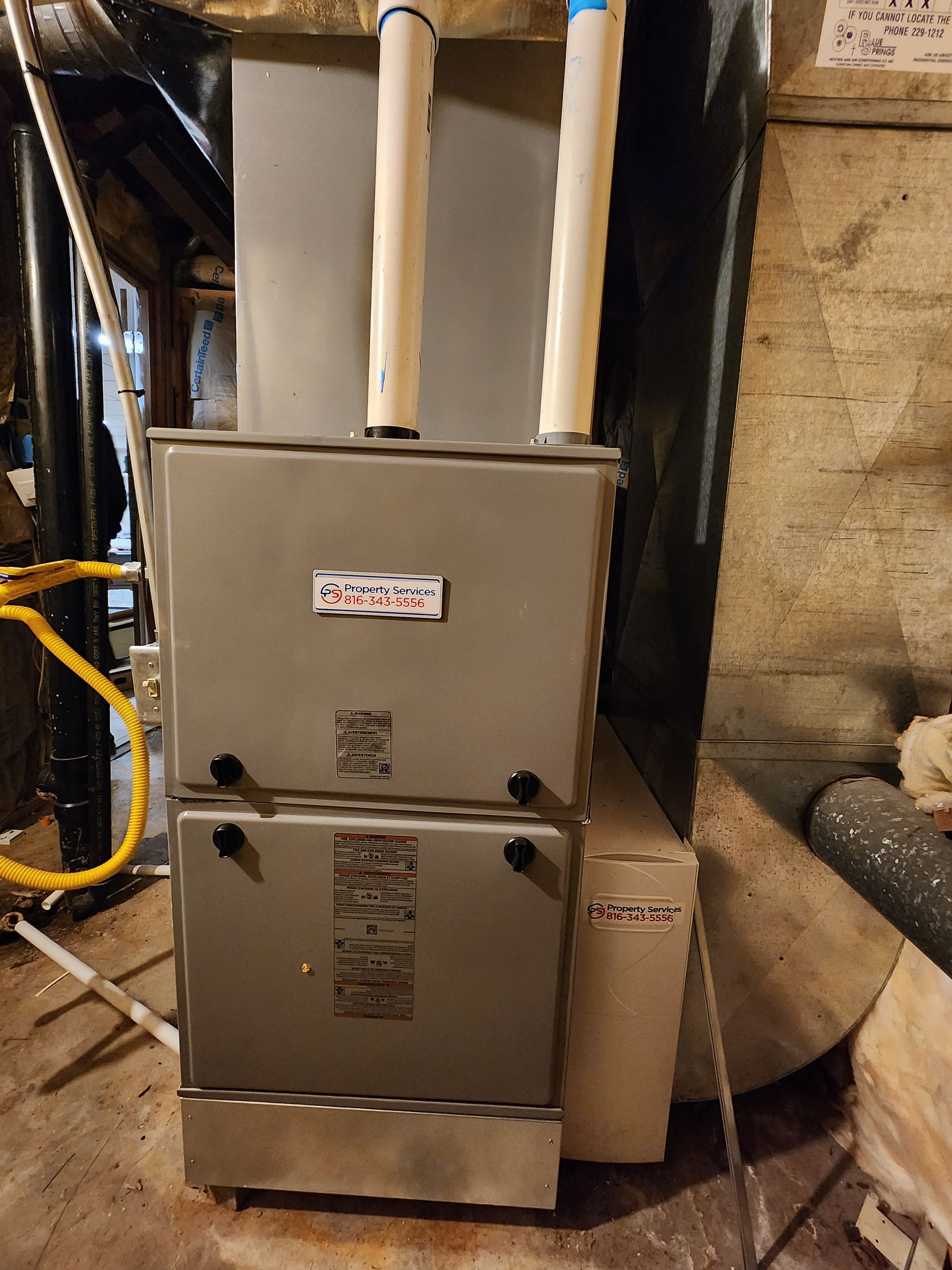 A gray high-efficiency residential furnace with two vertical white PVC vent pipes in a basement.