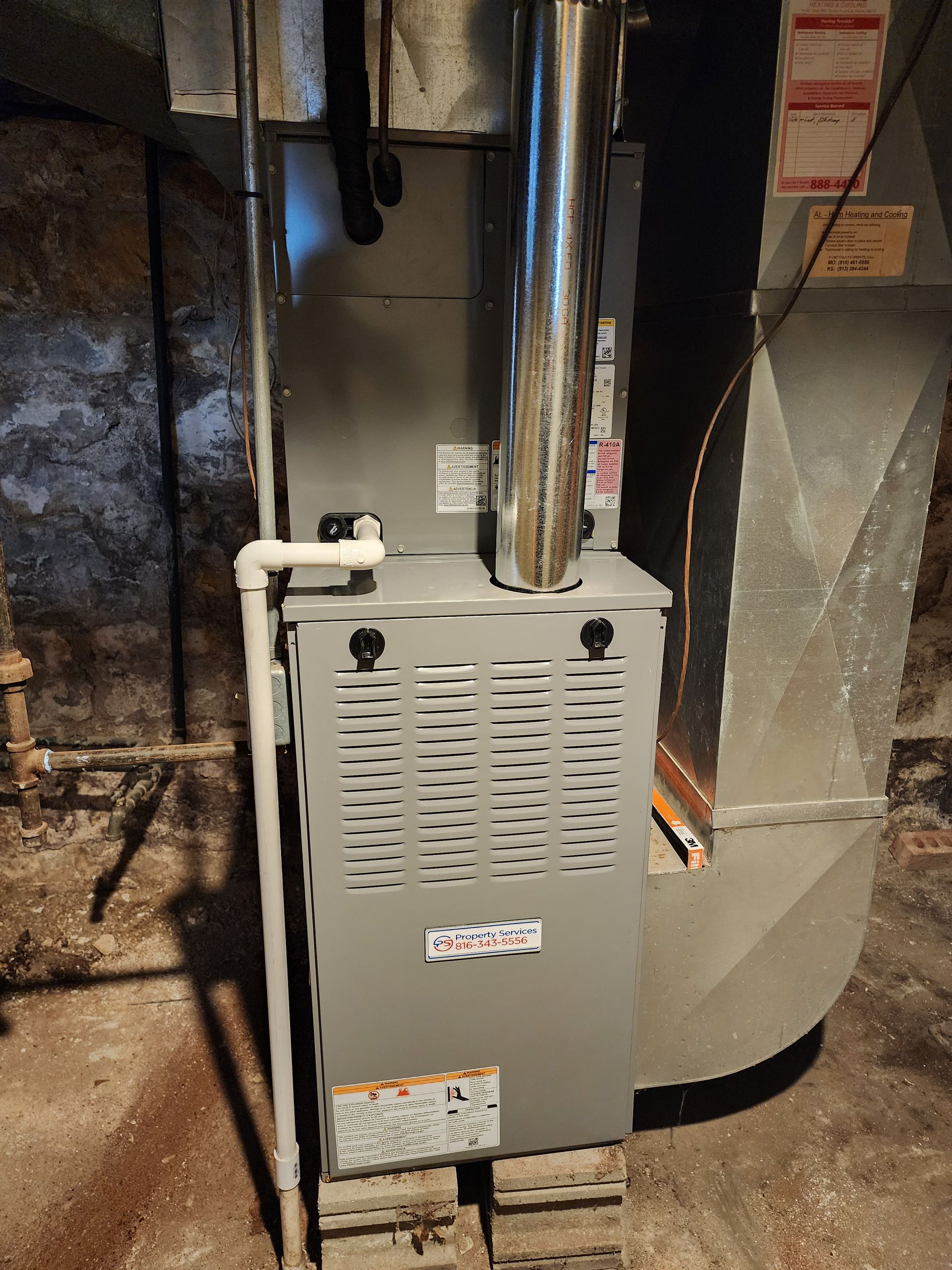 A gray HVAC furnace unit with a vertical silver flue pipe, positioned on concrete blocks in a basement setting.