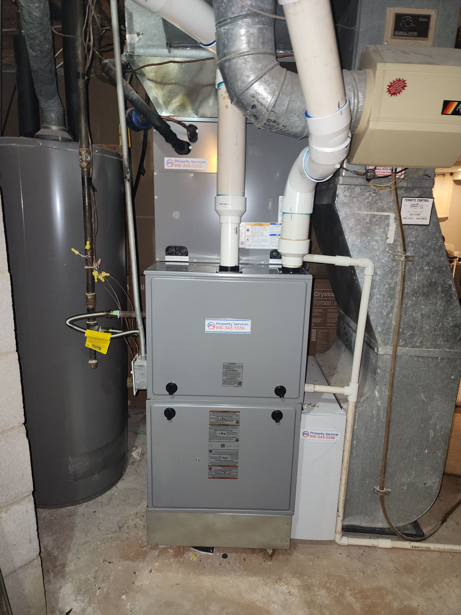 A gray HVAC furnace unit with white PVC intake and exhaust pipes installed in a basement next to a water heater.