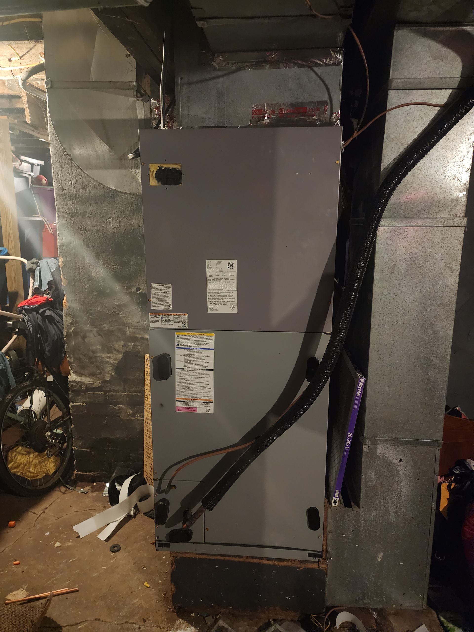 A gray HVAC air handler unit standing in a basement, connected to silver ductwork and insulated pipes.