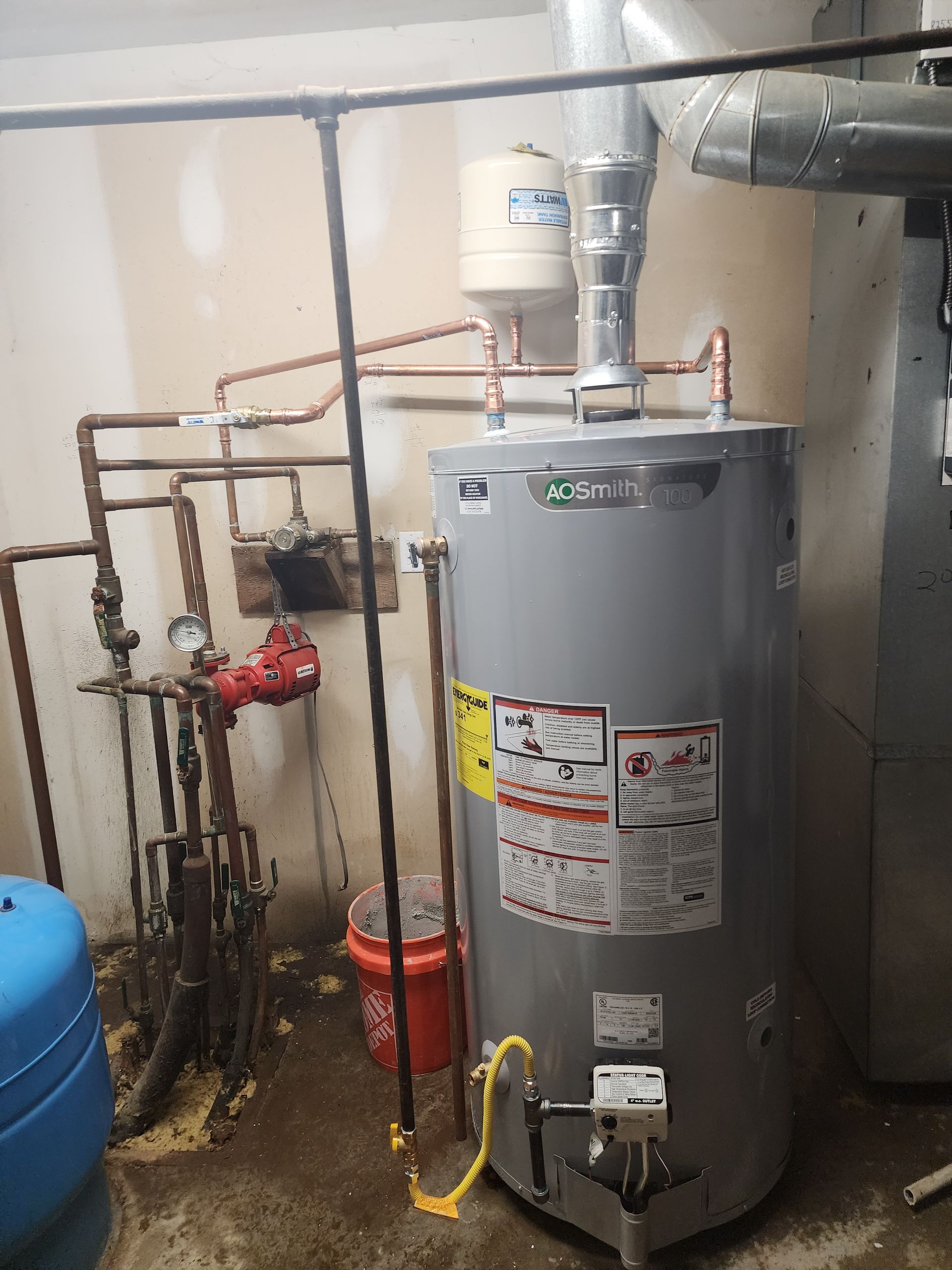A grey water heater in a utility room, connected to copper pipes, a vent, and a red pump with a bucket on the floor.