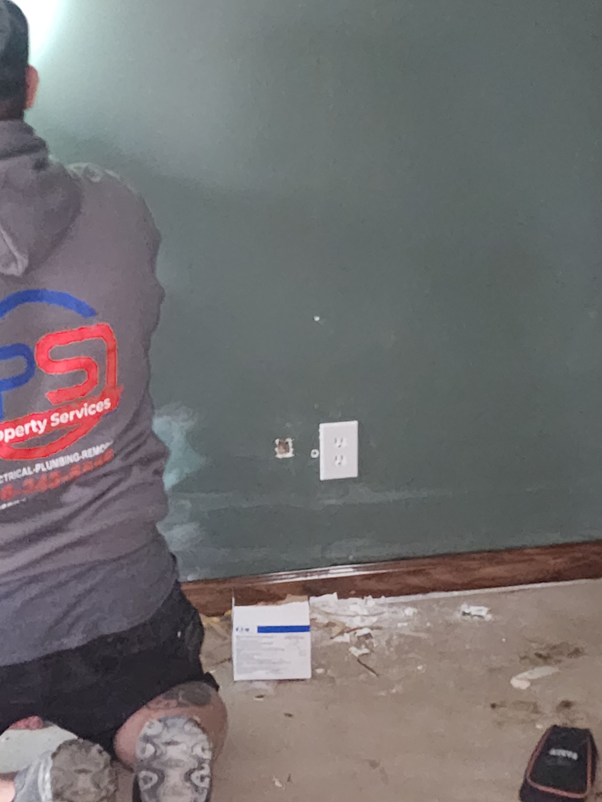 A person in a gray uniform kneels before a dark green wall, installing or repairing an electrical outlet cover.