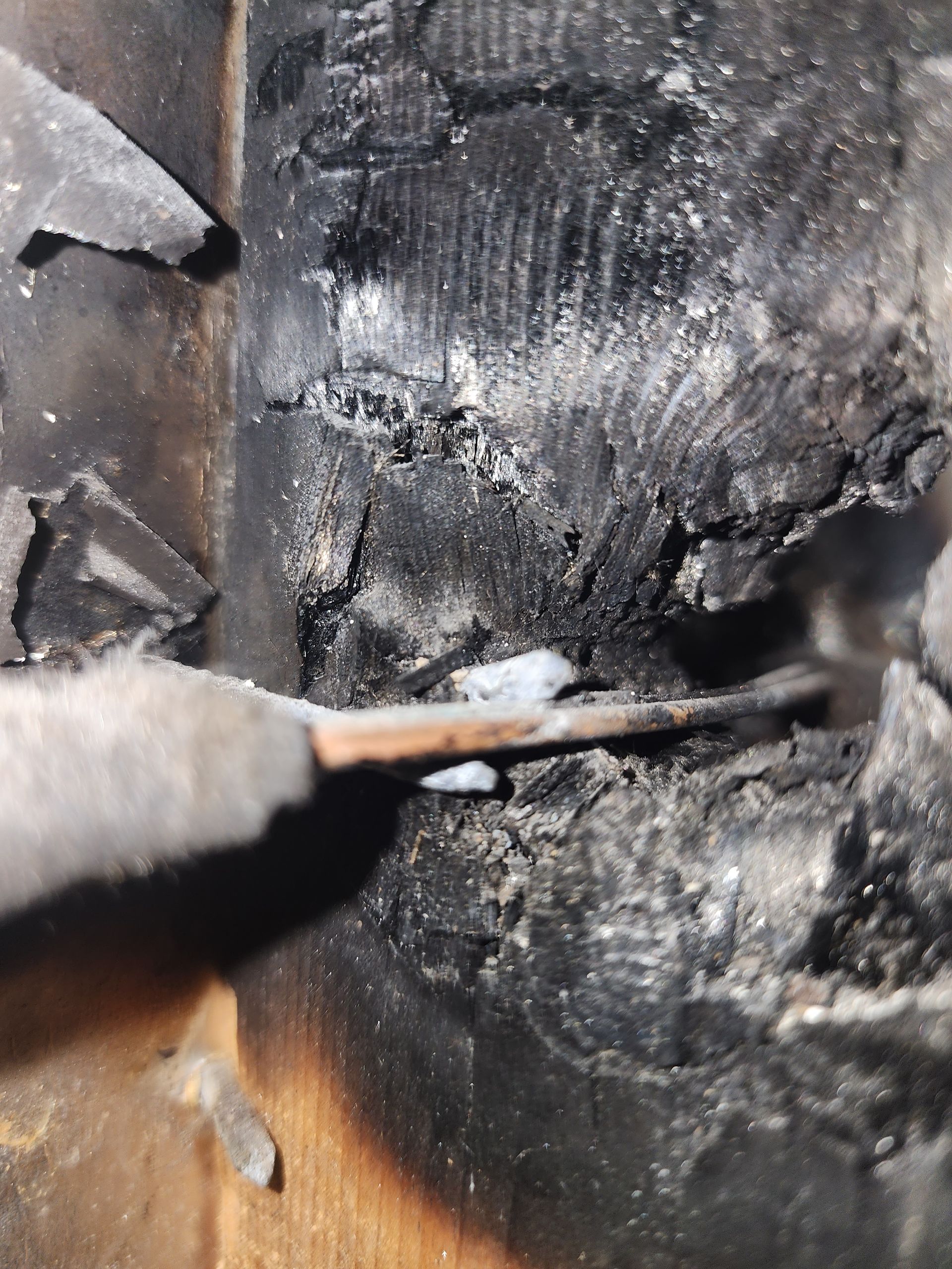 A close-up view of a metal tool probing a deep crack in charred, black, textured wood.