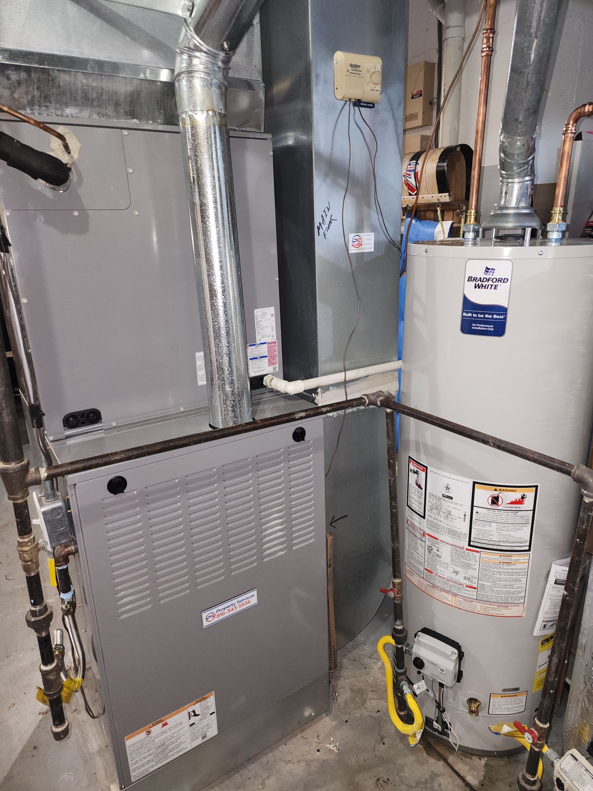 A gray furnace and a light-colored water heater stand side-by-side in a utility area with exposed pipes and metal vents.