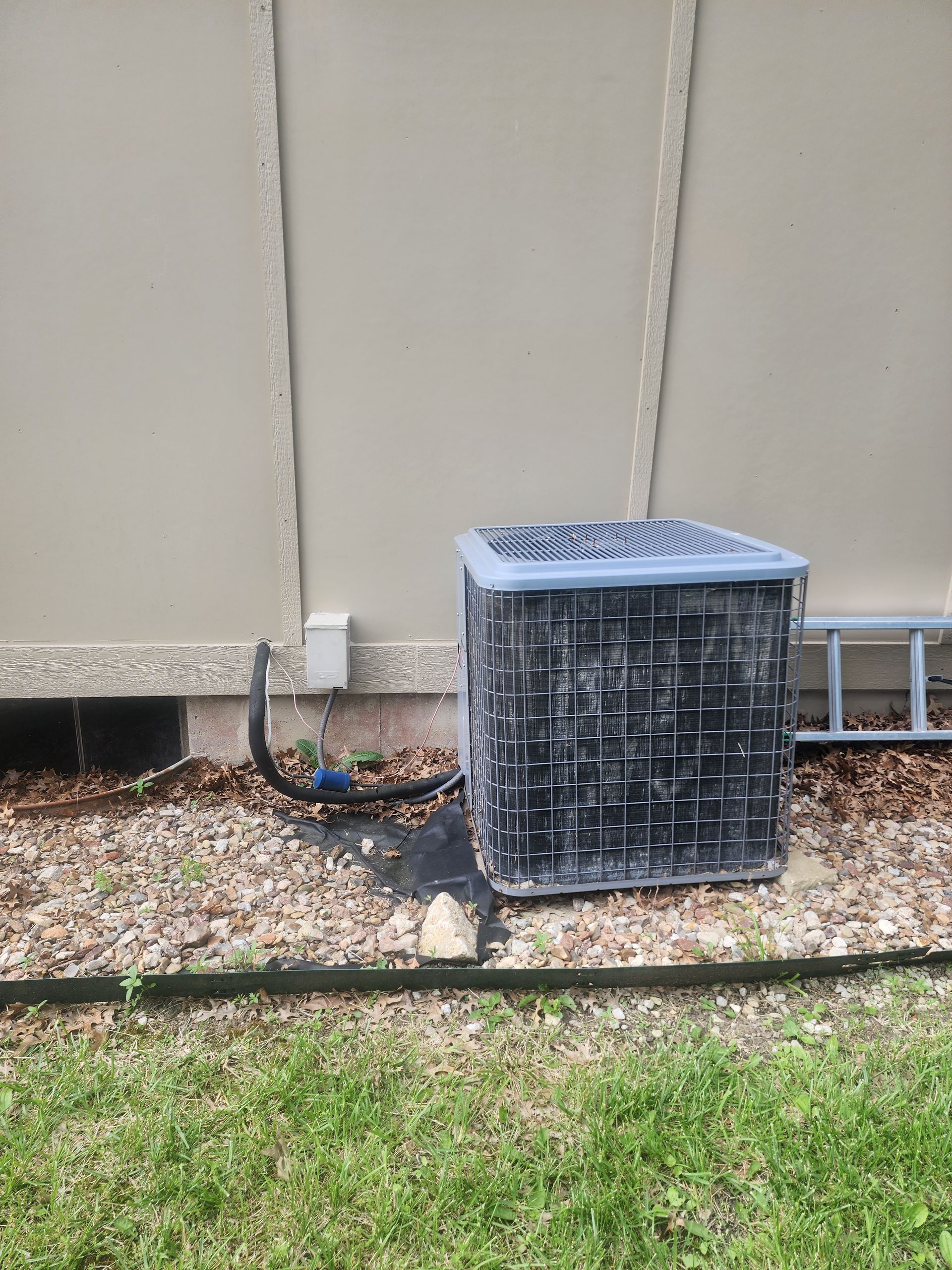 A gray residential air conditioning unit sits outdoors on a gravel bed next to a tan house wall.