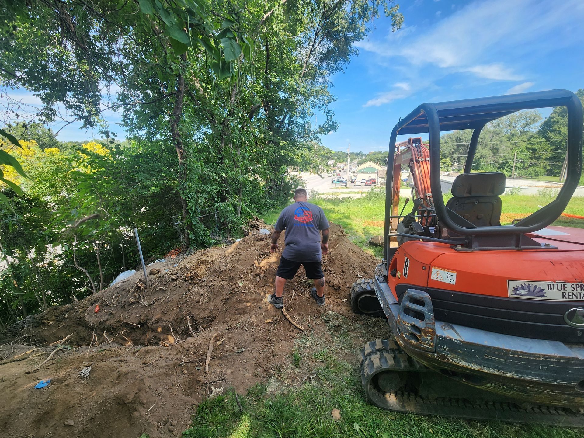 A person in a gray shirt works on a pile of dirt next to an orange excavator in a grassy, wooded area on a sunny day.