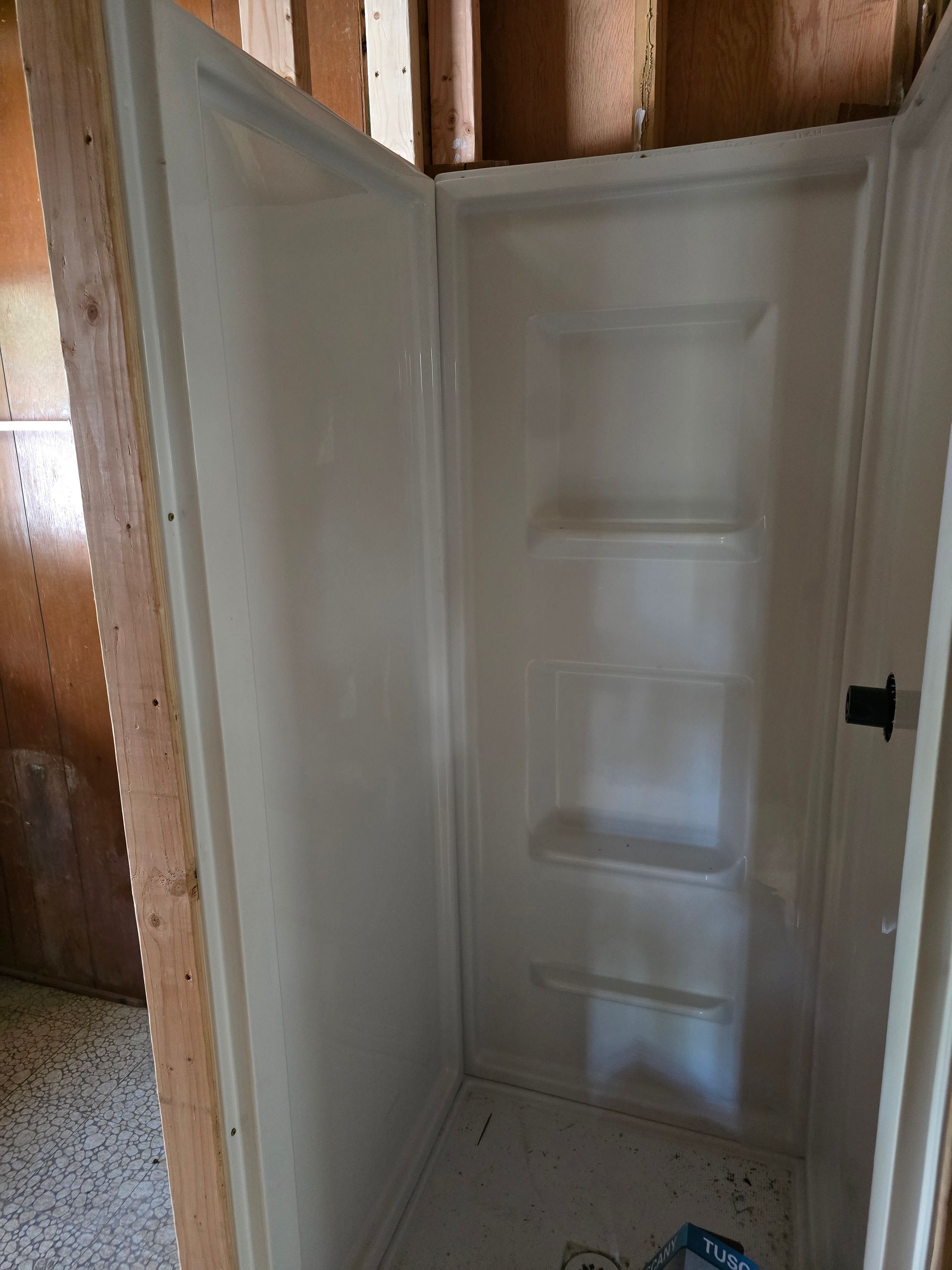 A white, pre-fabricated shower stall with built-in shelving, installed in an unfinished room with wooden wall studs.
