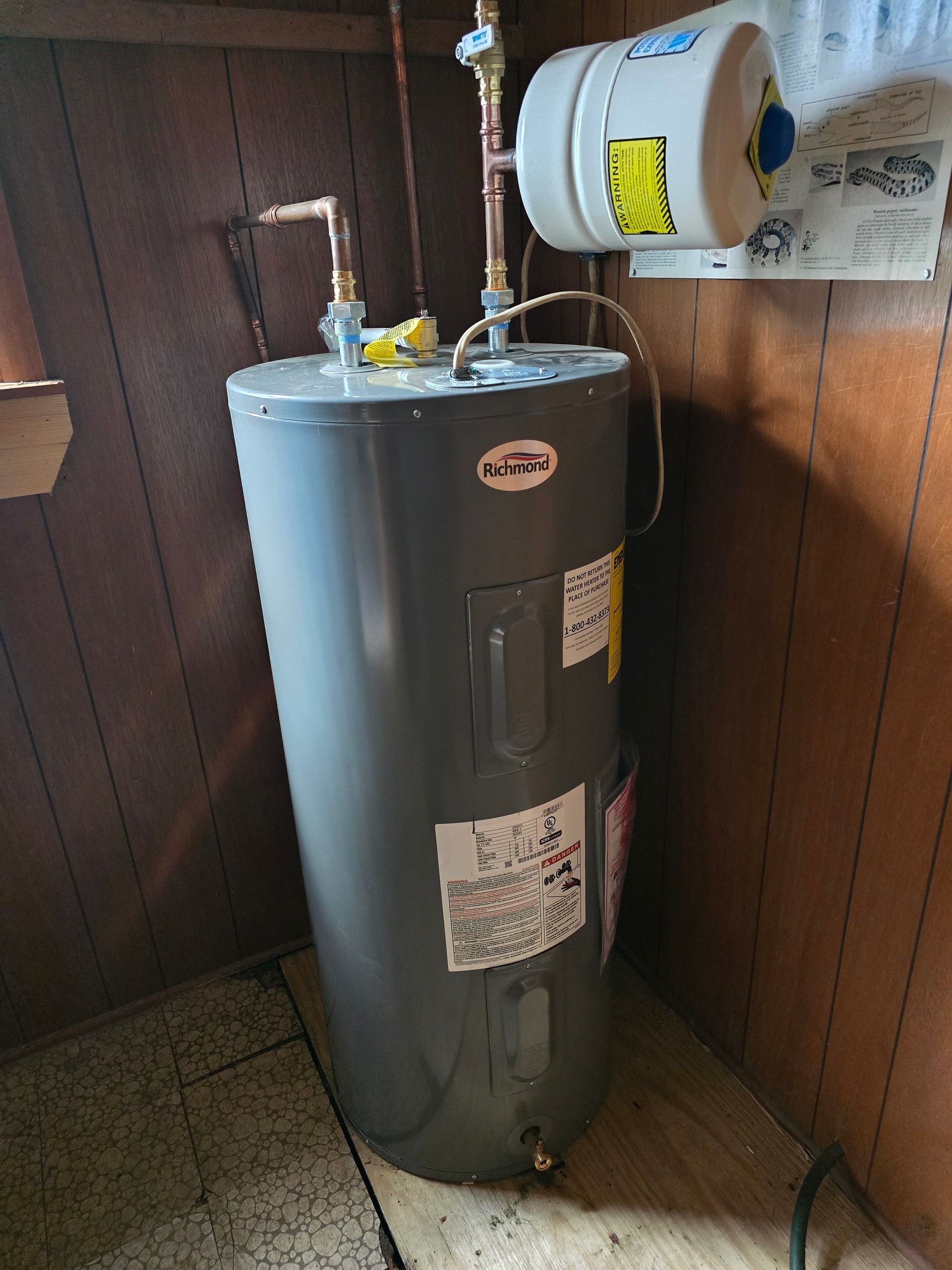 A gray electric water heater with an attached thermal expansion tank stands in a corner with wood-paneled walls.