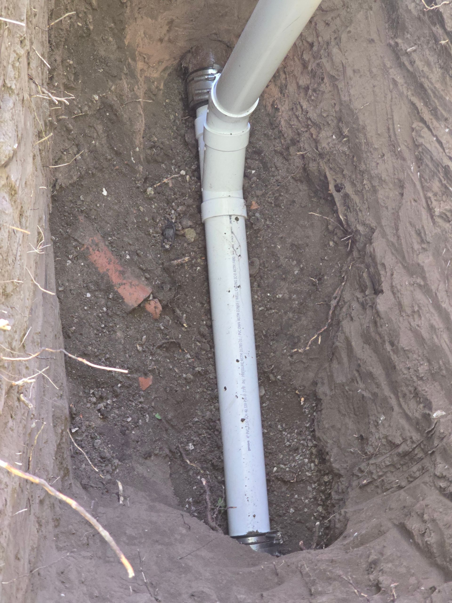 A white PVC pipe connected to a metal pipe in a narrow, dirt-filled trench.