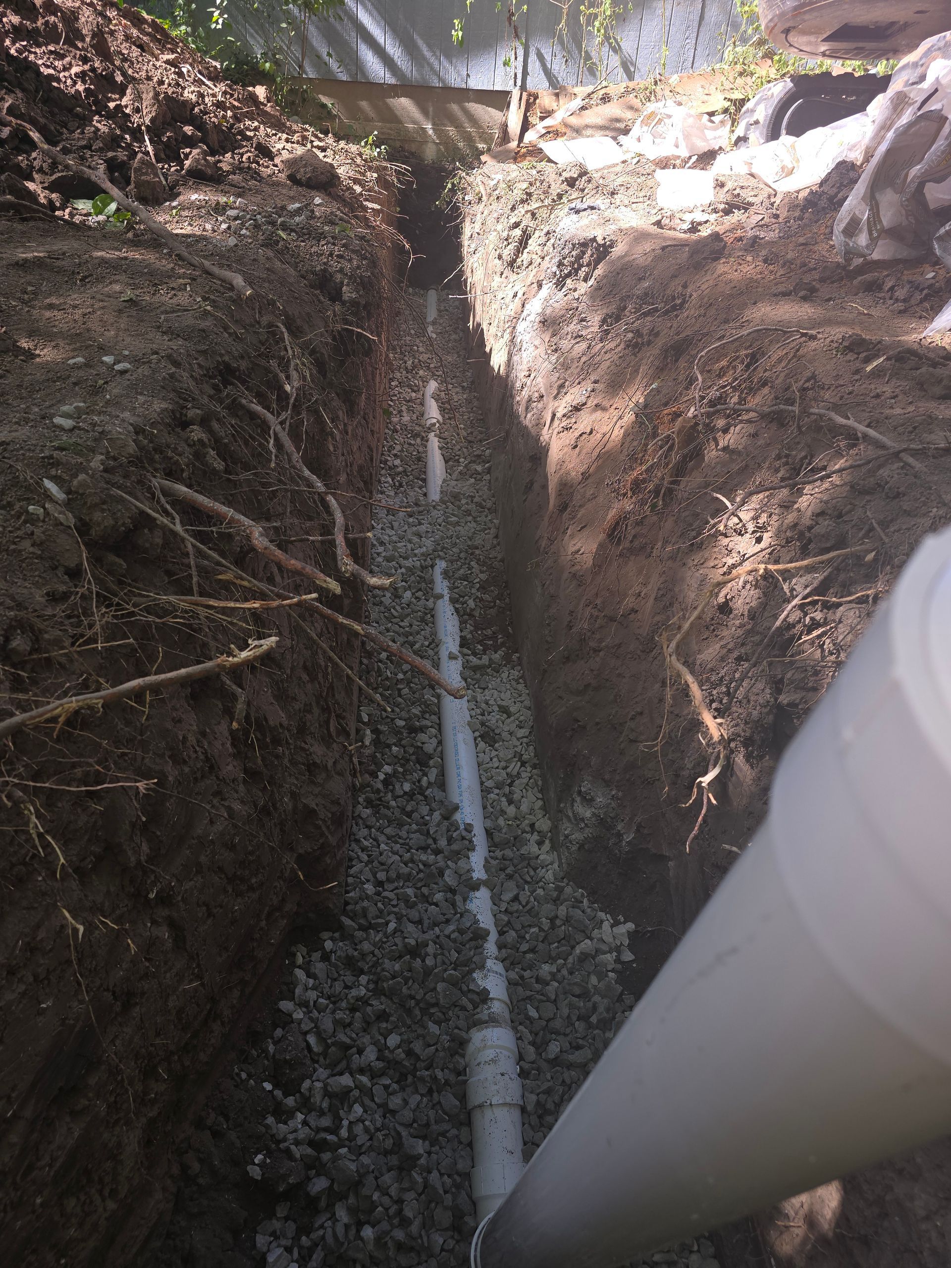 A white PVC drainage pipe lying in a narrow dirt trench filled with gravel, viewed from above.