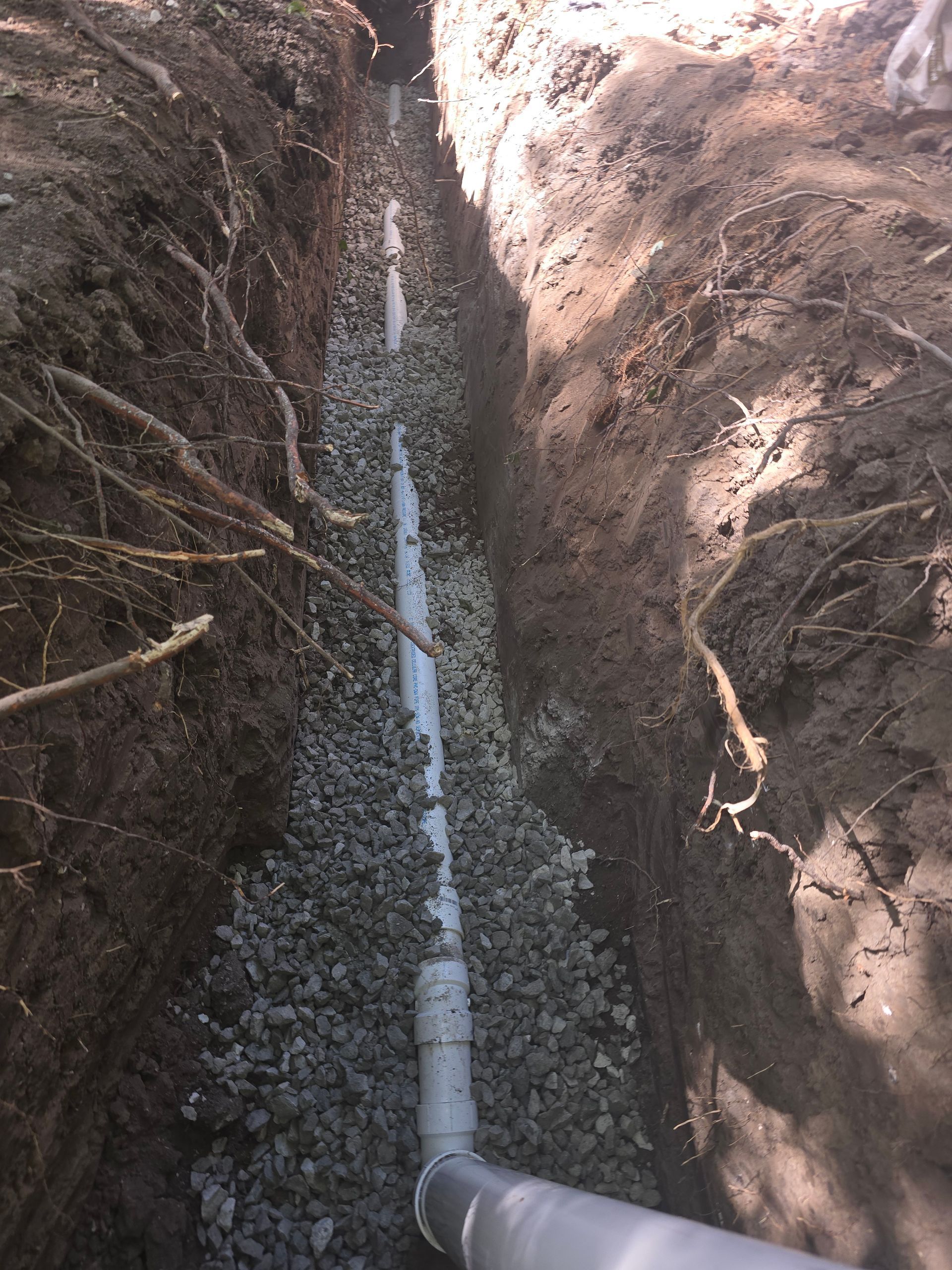 A white PVC drainage pipe installed in a narrow trench surrounded by gravel and dark soil.