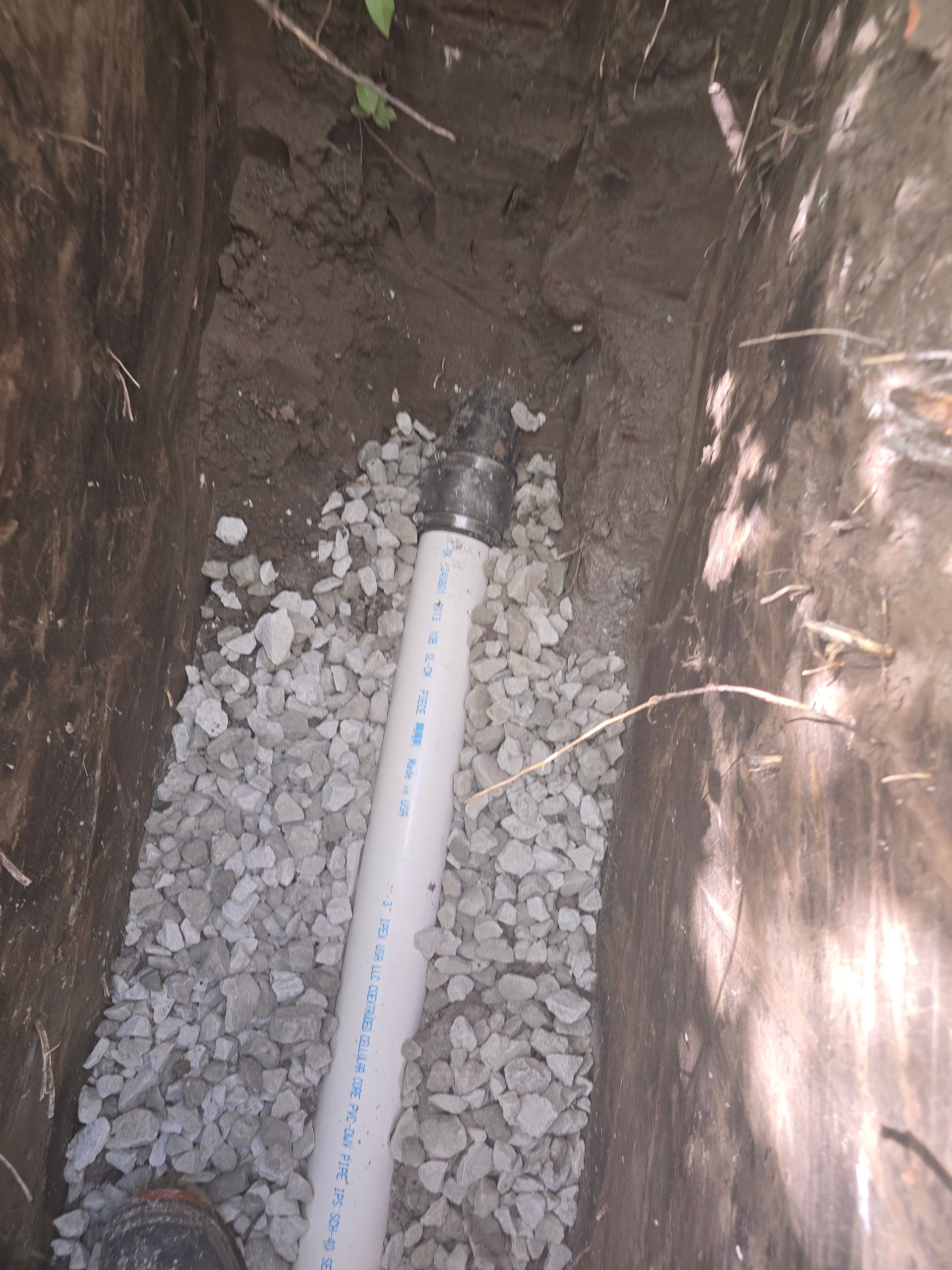 A white PVC pipe runs through a narrow trench, with a metal coupling and gravel base.