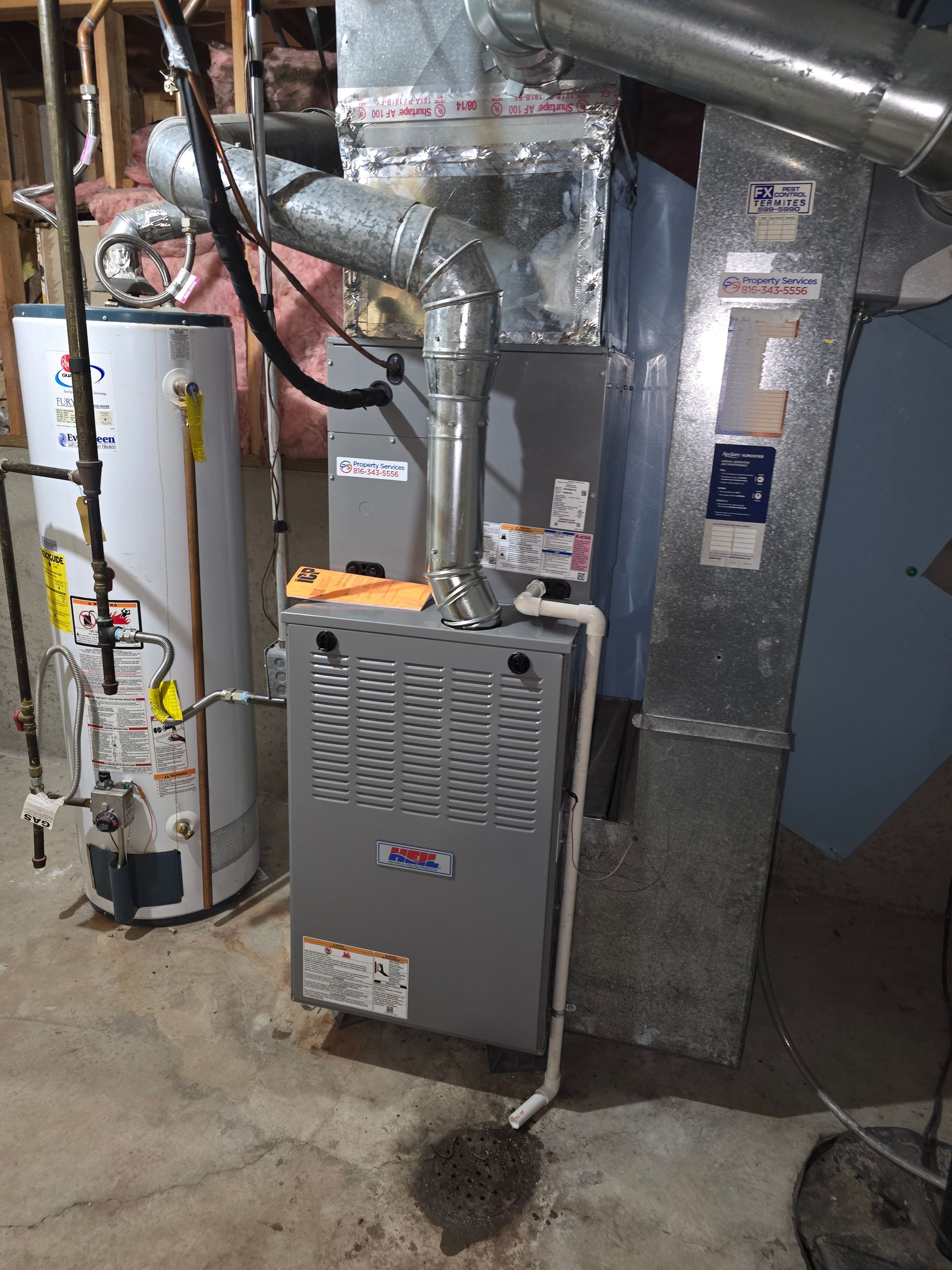 A white water heater stands beside a gray HVAC furnace unit in a basement, with metal ductwork and piping overhead.