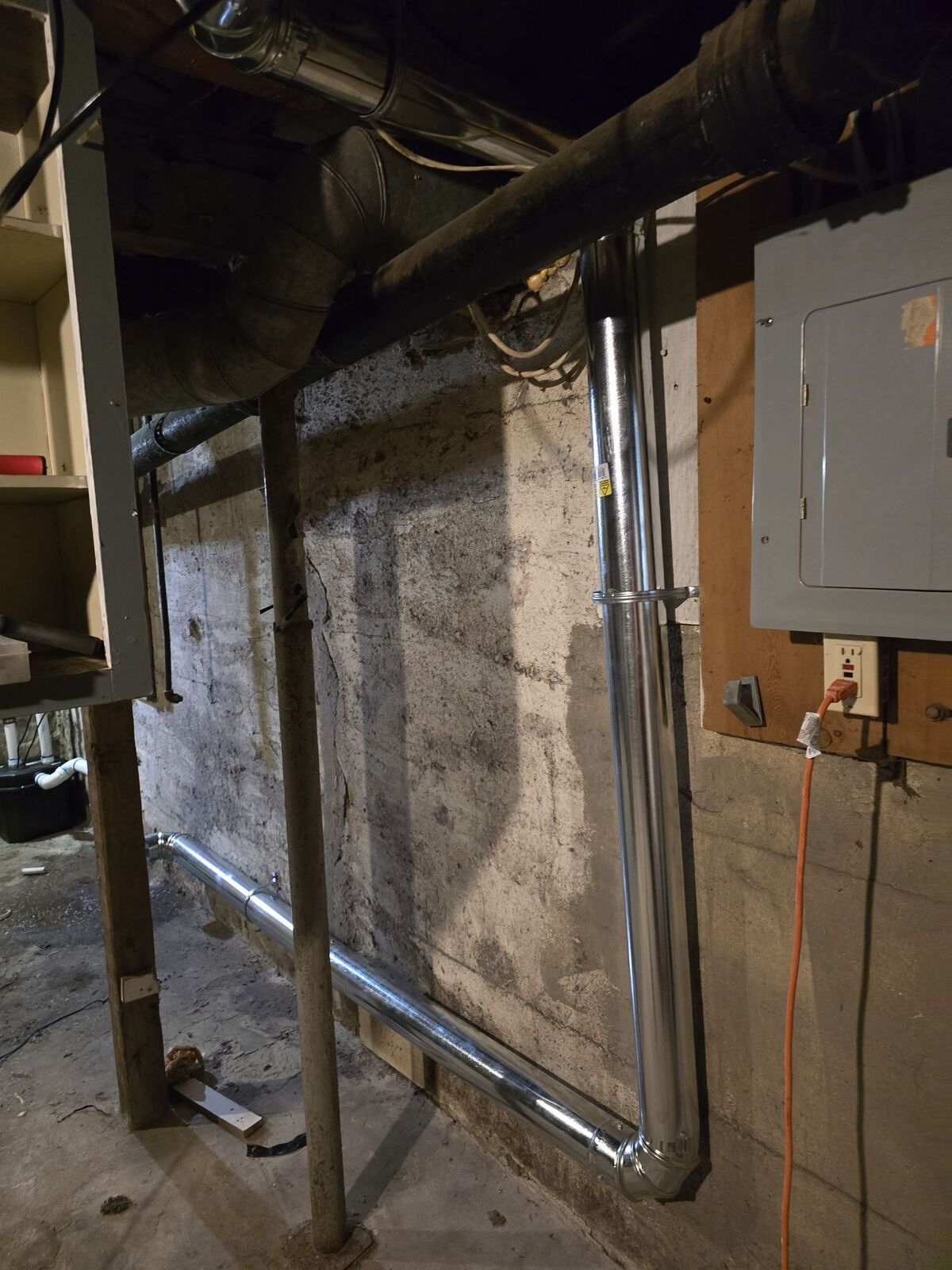 A metal duct runs down a concrete basement wall and turns at a right angle along the floor near a wooden electrical panel.
