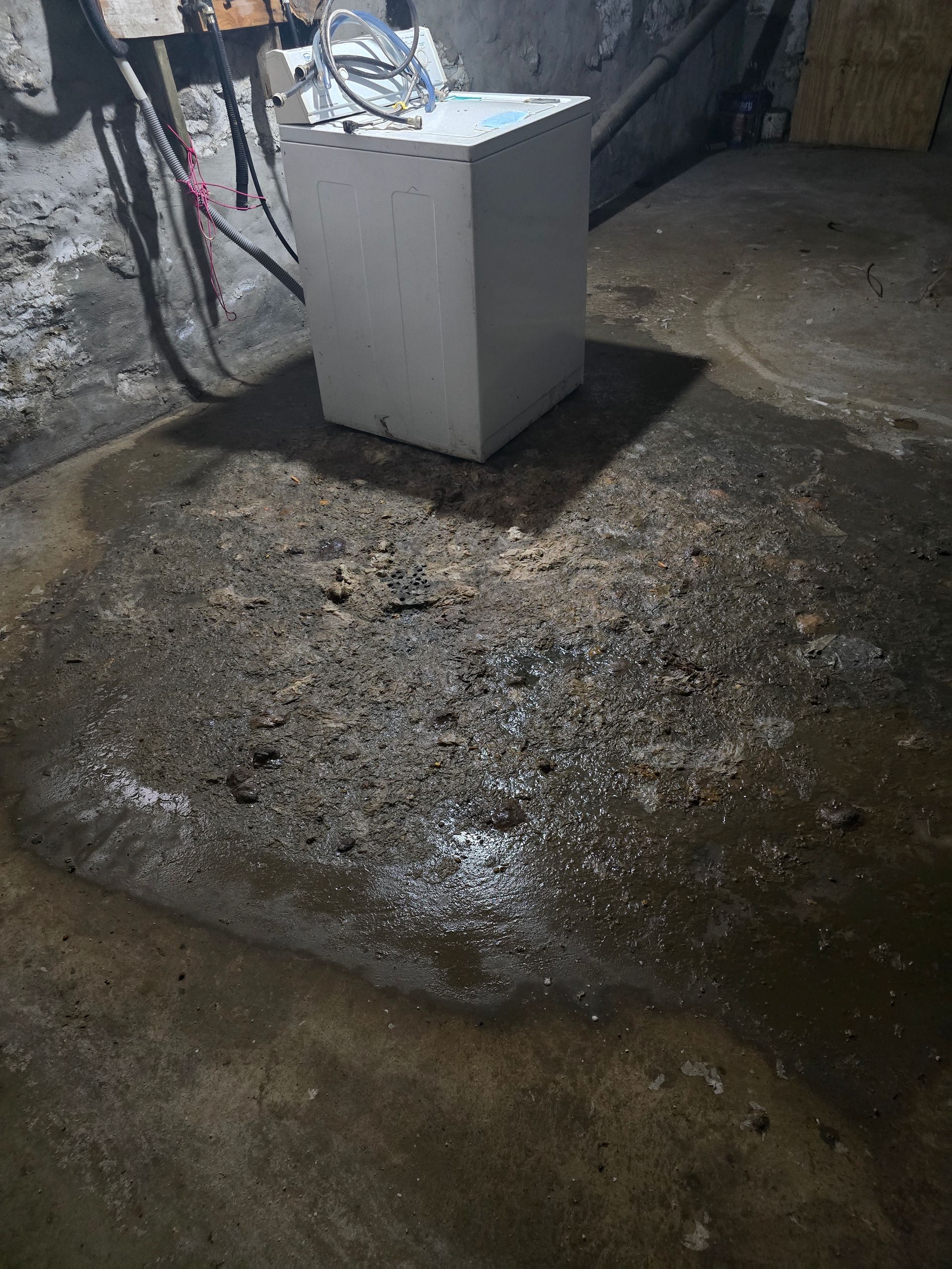 A white appliance sits on a concrete floor in a basement with a patch of damp, uneven, and crumbling ground in front of it.