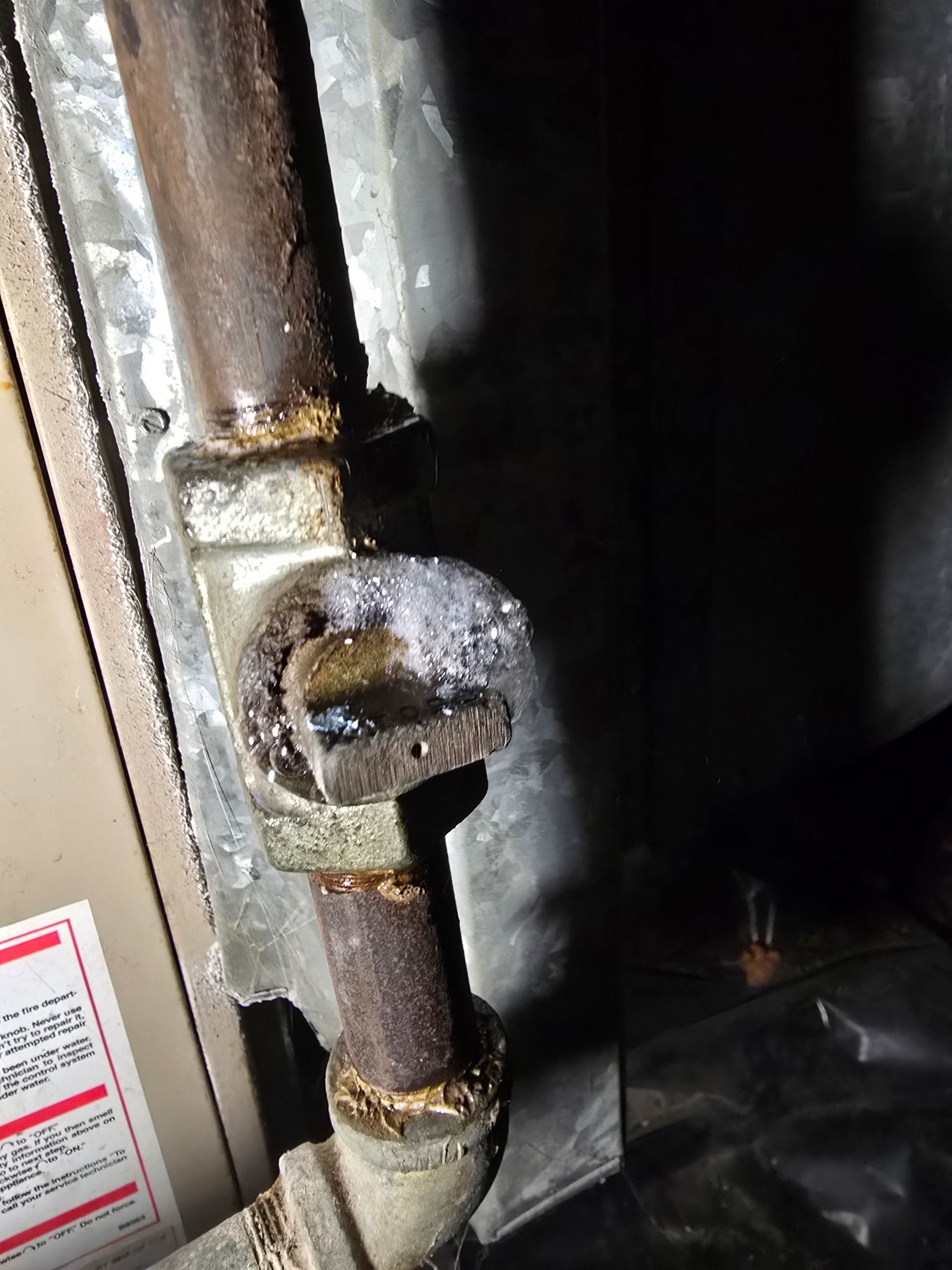A rusty metal gas valve on a pipe attached to a furnace, covered with a patch of white frost or ice.