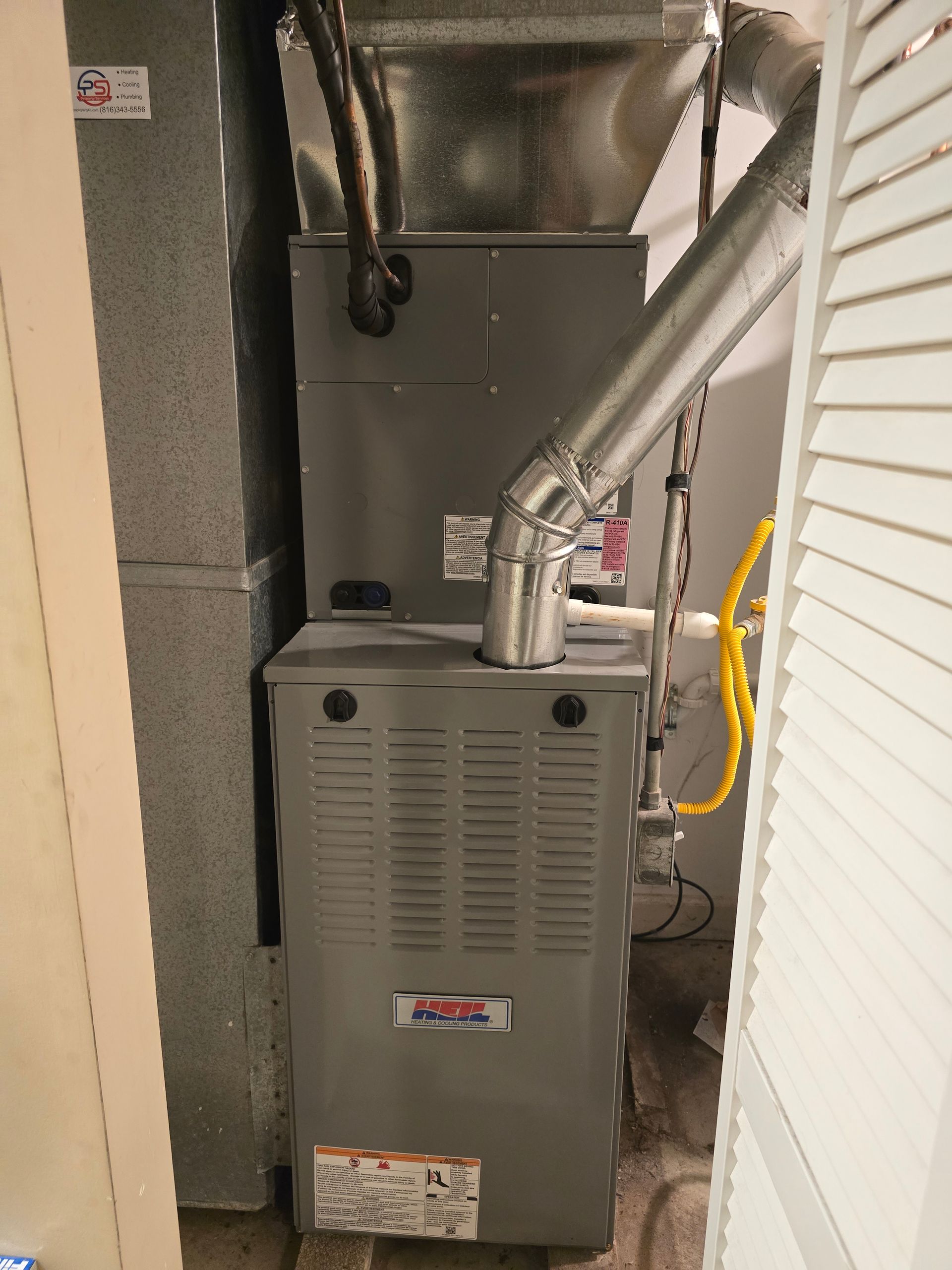 A gray, upright HVAC furnace unit with metal venting, located in a narrow utility closet.