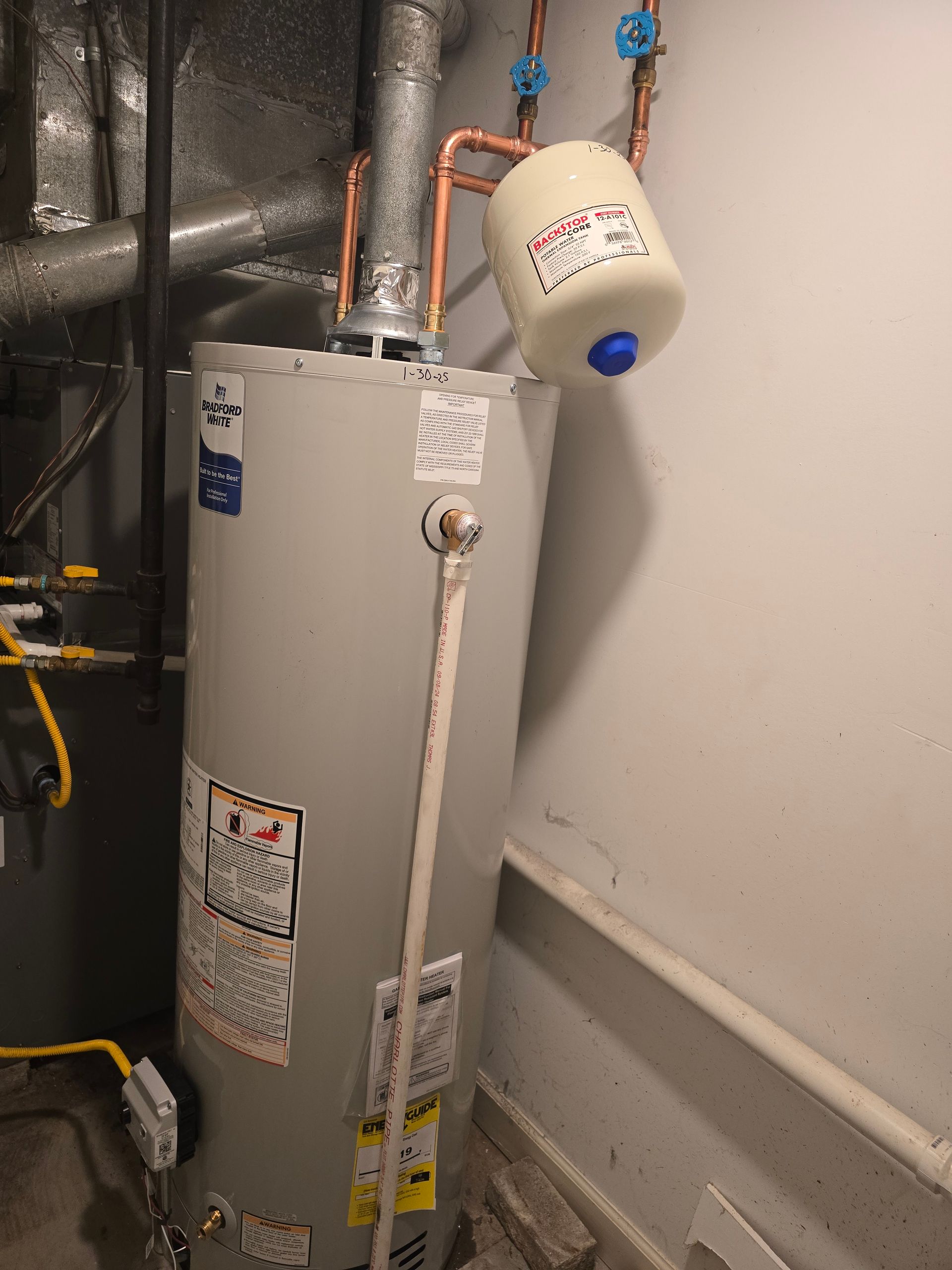 A gray residential water heater with a beige expansion tank mounted on copper pipes above it in a utility space.