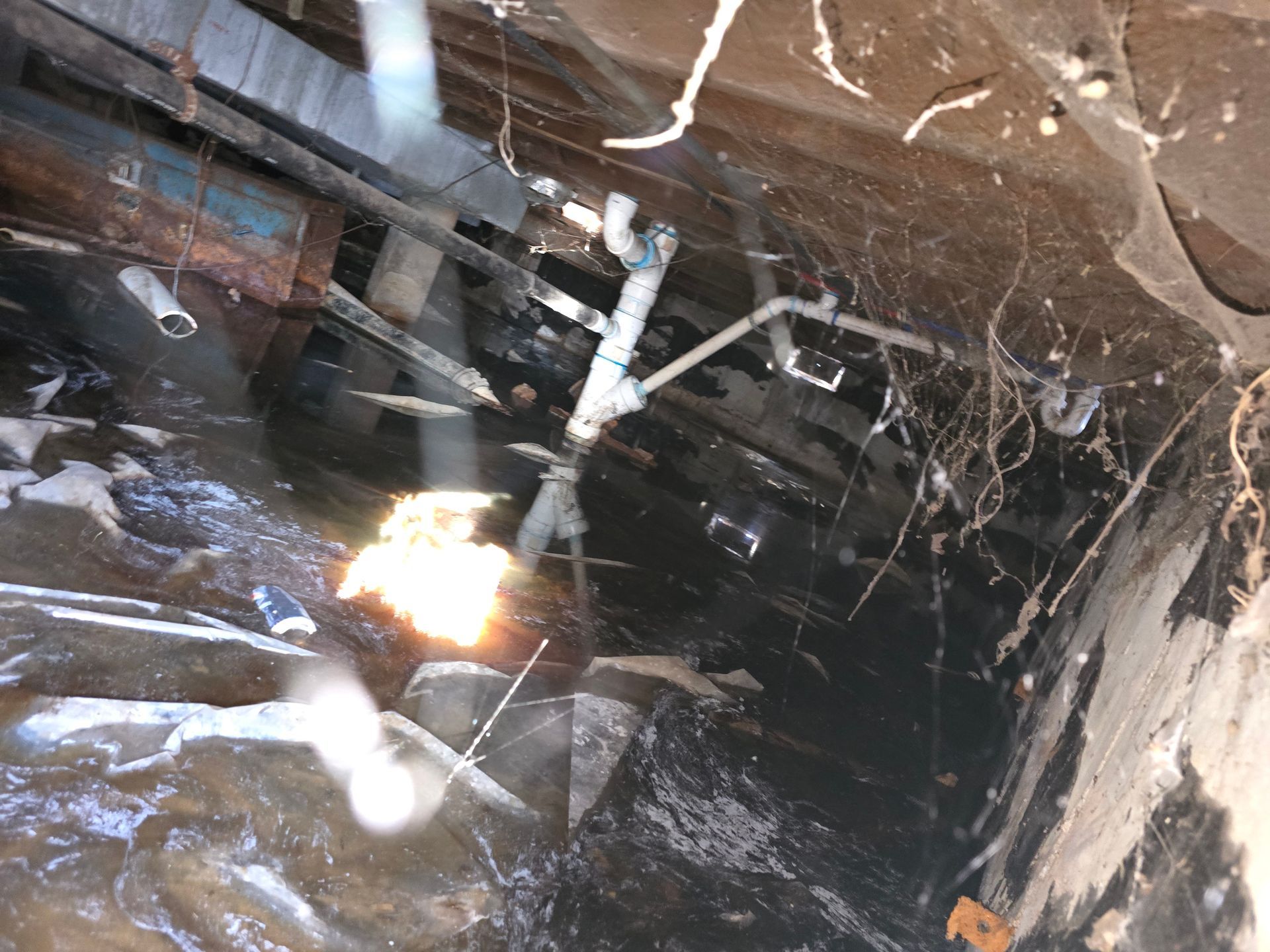 A dark, damp crawl space with exposed pipes, mud-covered ground, and debris under a building foundation.