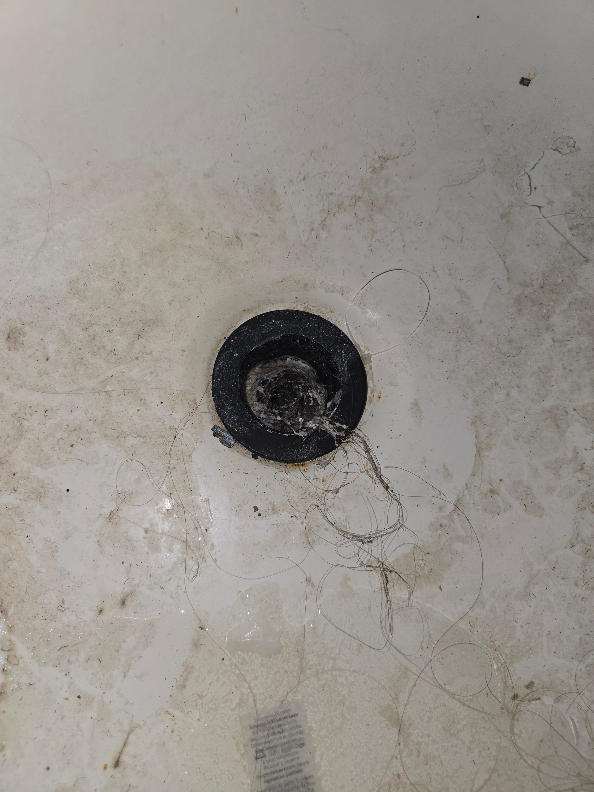 A dirty white shower floor with a dark drain surrounded by hair and debris.
