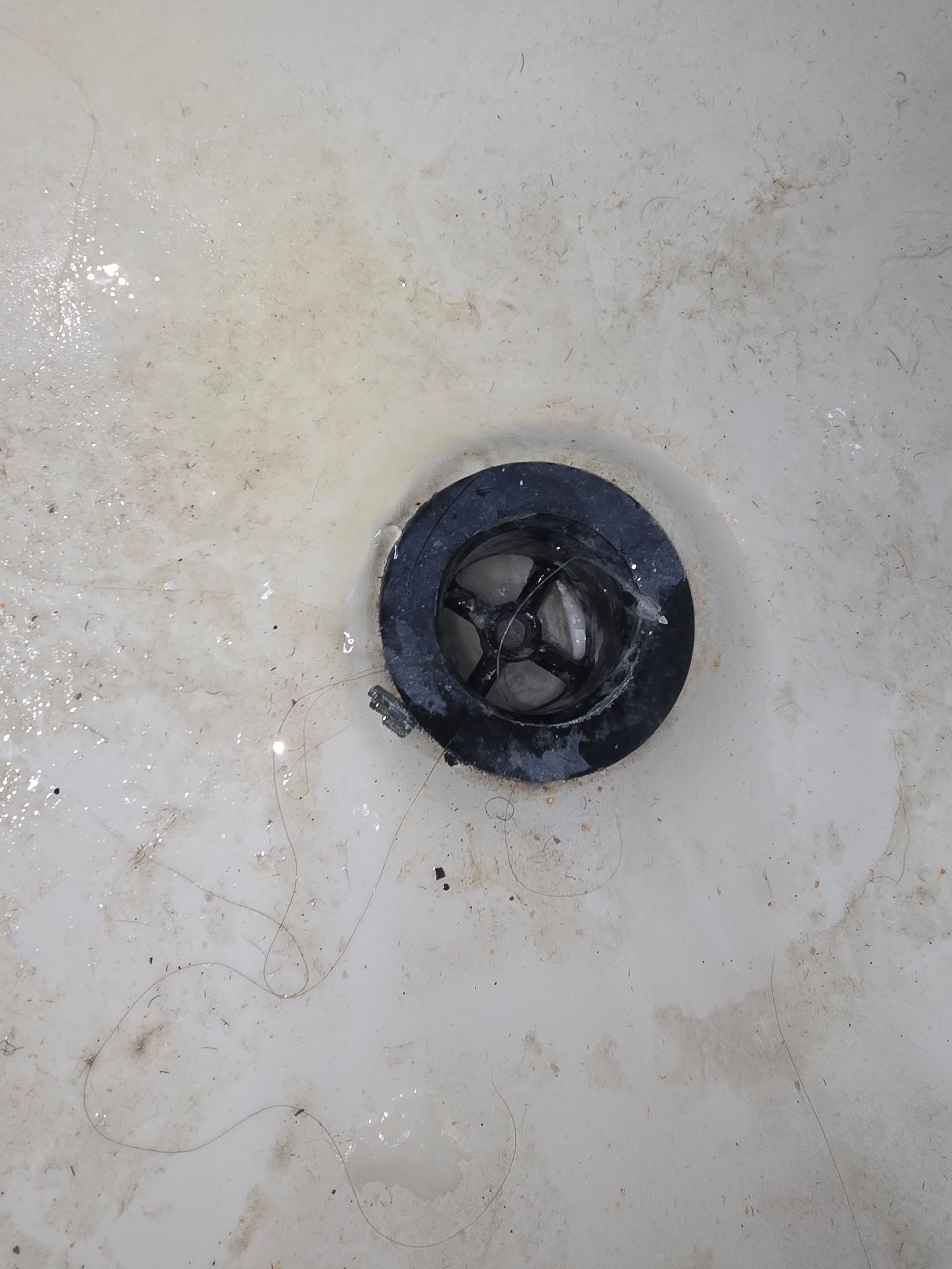 A close-up of a dirty, light-colored bathroom sink drain with a black rubber hair catcher insert.