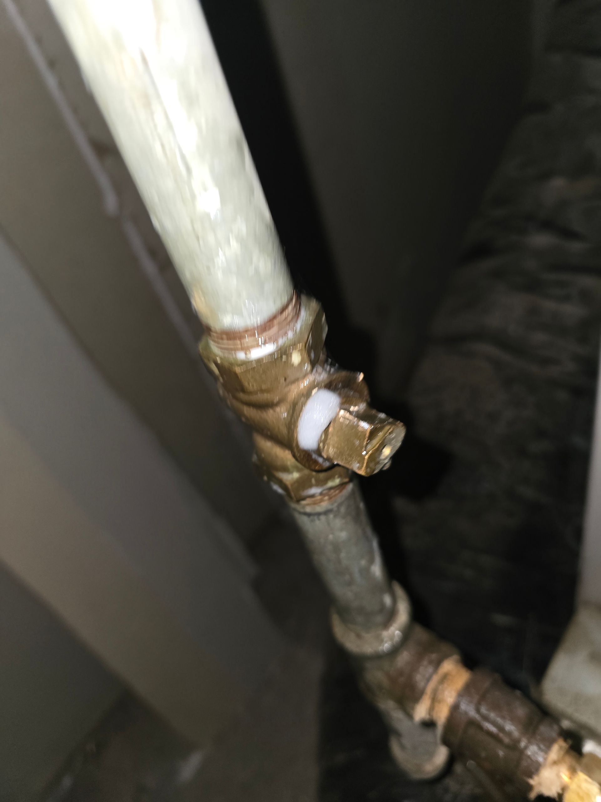 A brass pipe valve connector with a white plastic seal, installed on a metal pipe system in a dimly lit area.