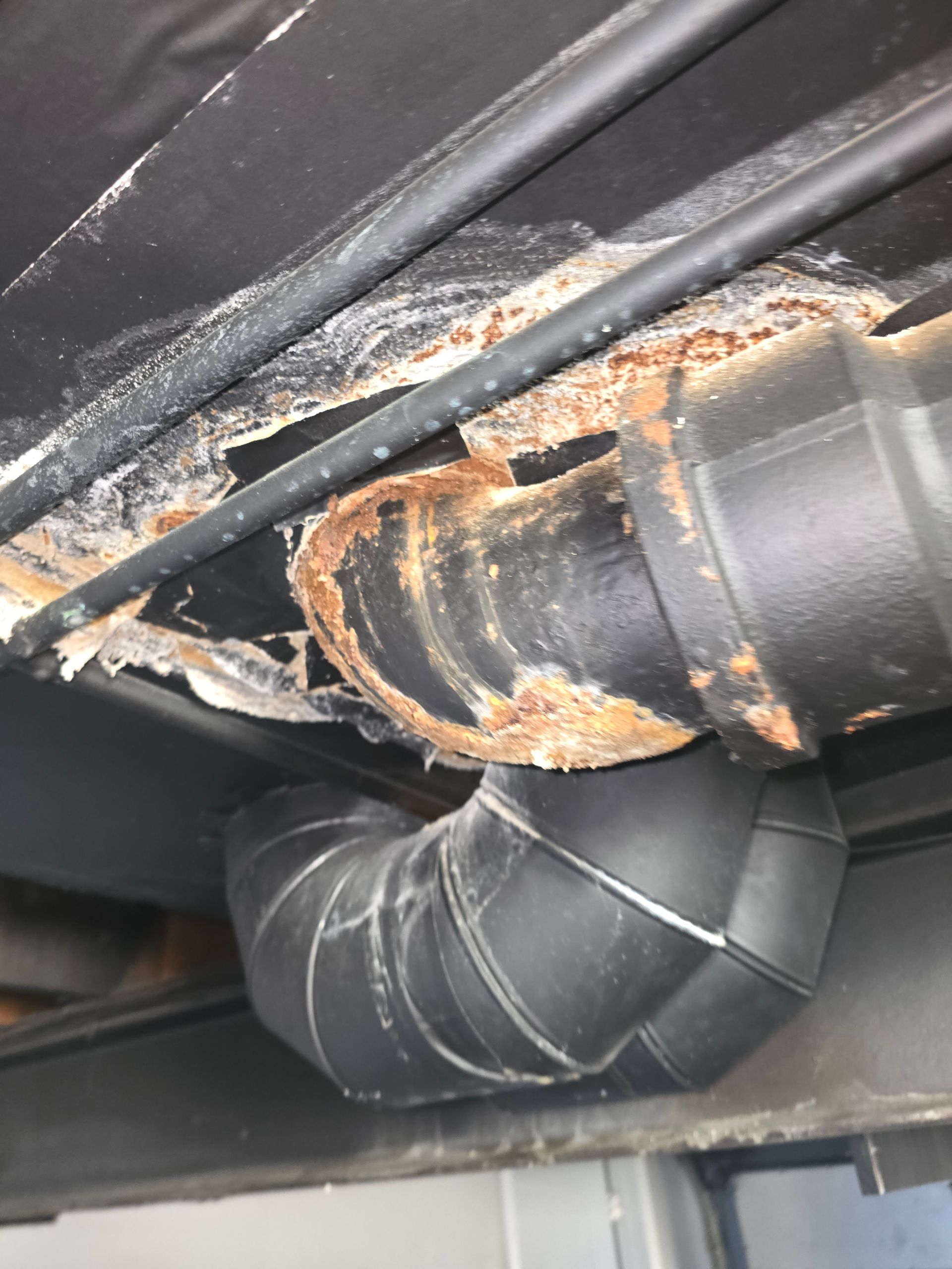 Corroded black plumbing pipe showing significant rust and structural decay, hanging beneath a dark ceiling surface.