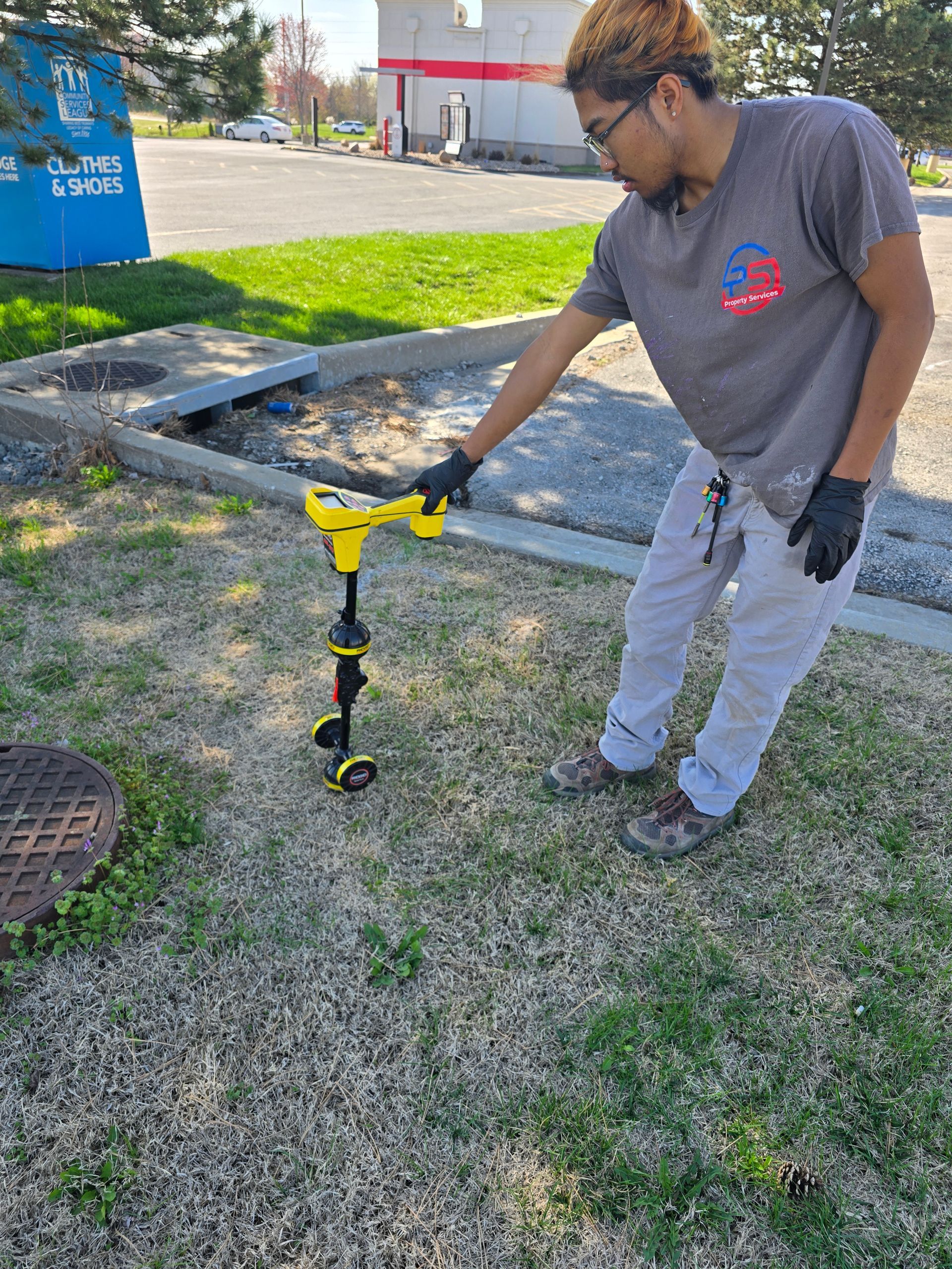 A person in a gray shirt and black gloves uses a yellow handheld earth auger to drill into the grass near a parking lot.