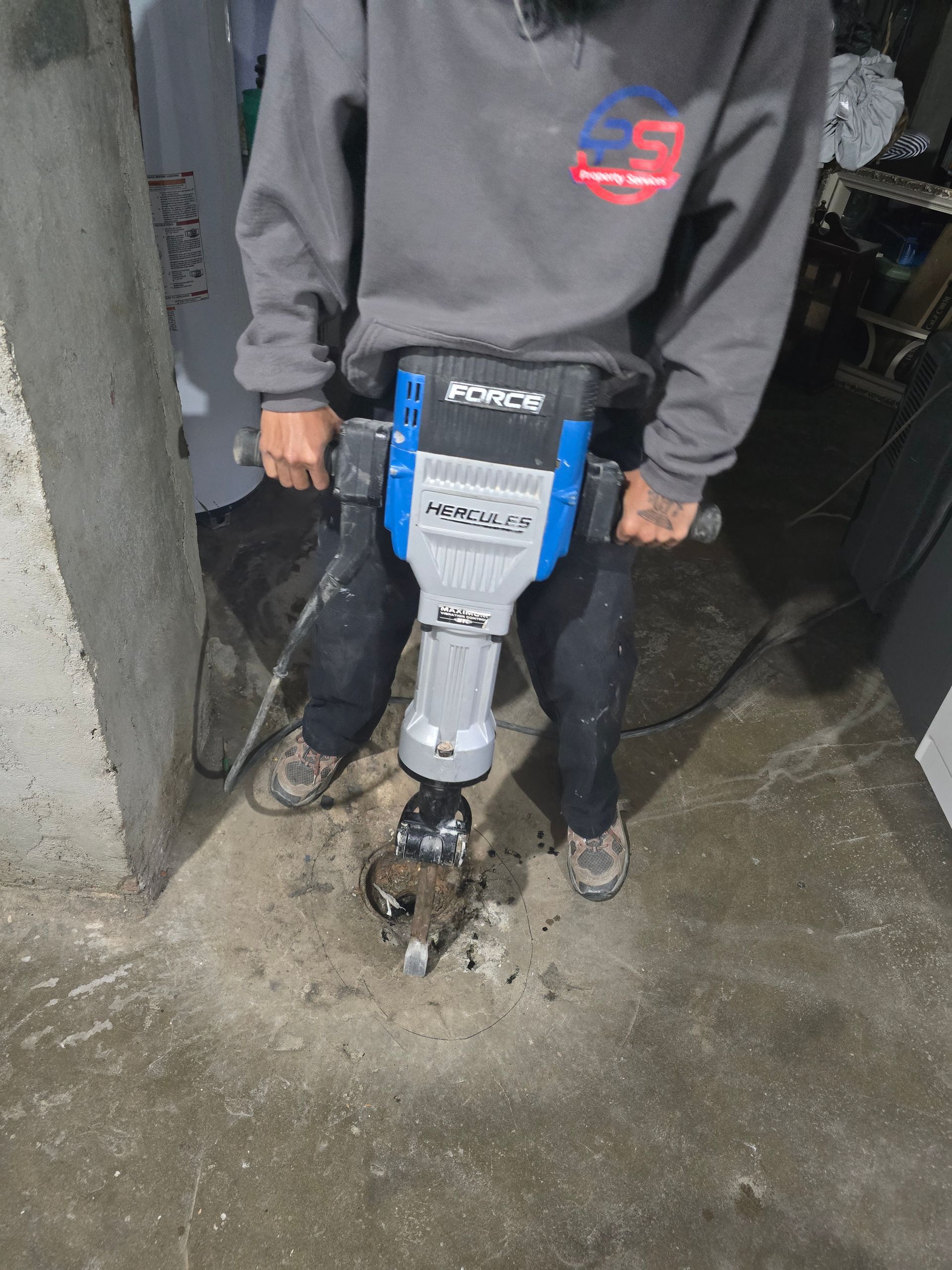 A person uses a blue and grey electric jackhammer to break up concrete on a floor.