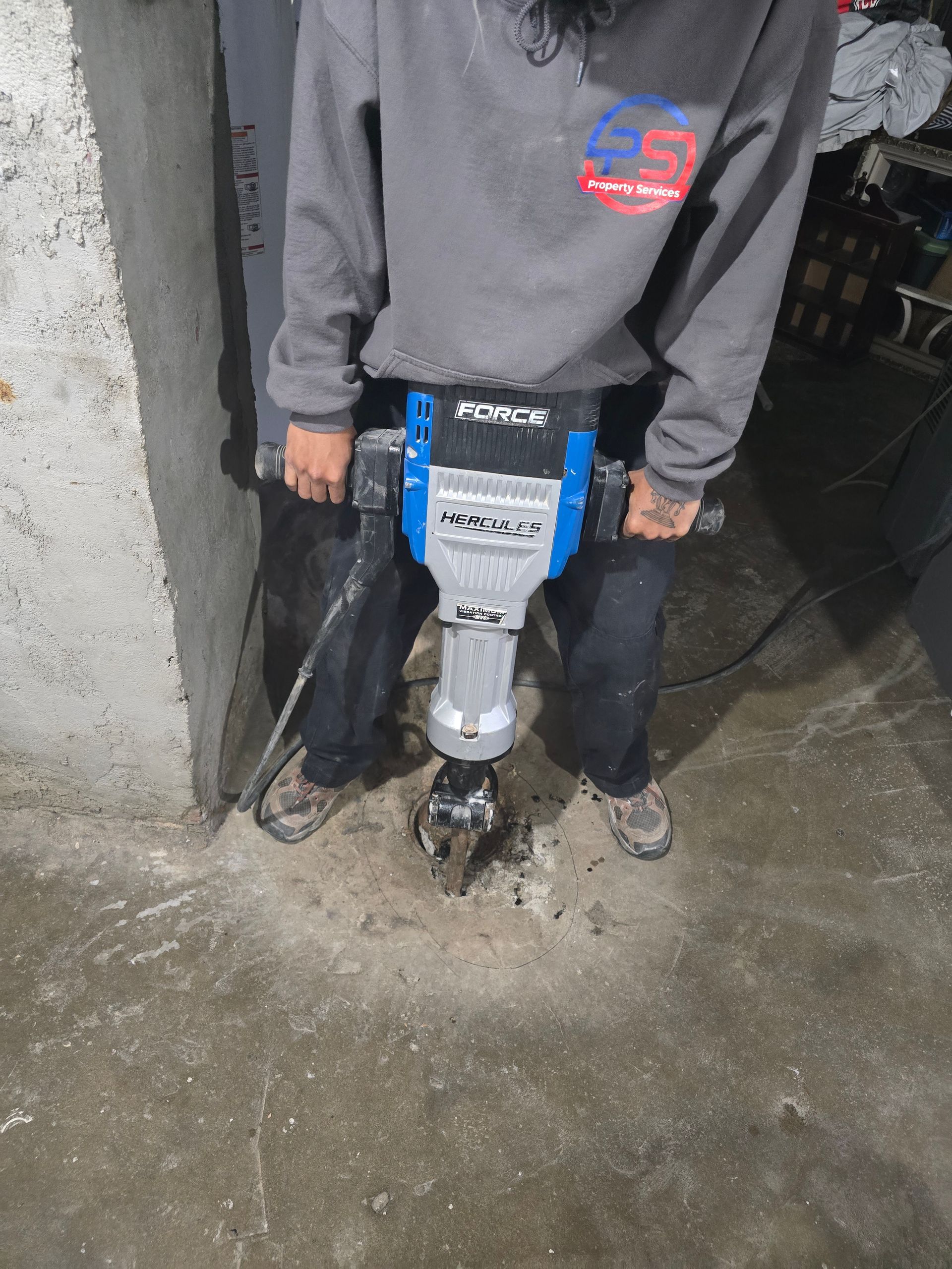 A person in a grey sweatshirt uses a blue and silver electric jackhammer to break through a concrete floor.